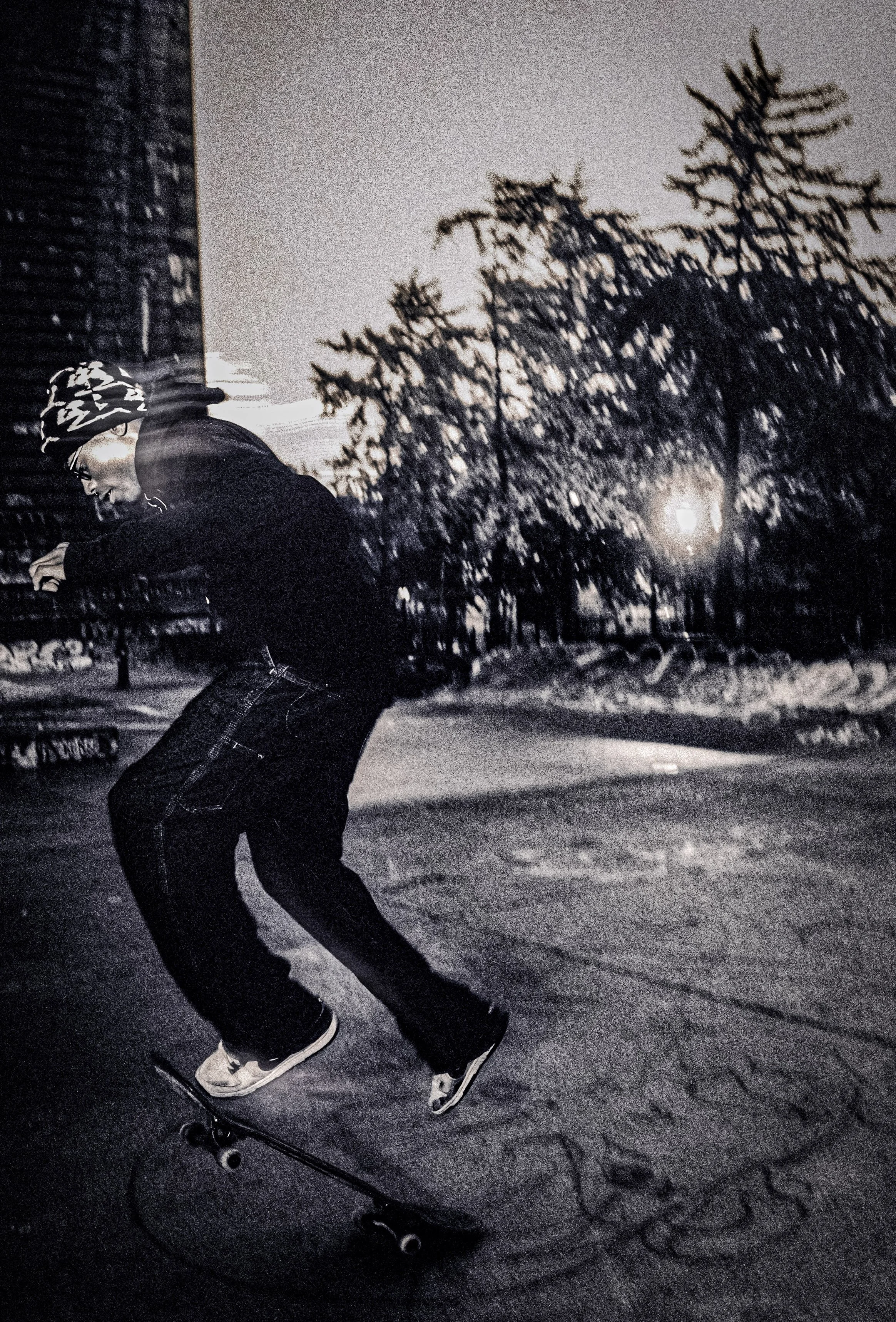 A person skateboarding outdoors at night, wearing a camouflage beanie and glasses, with trees and a streetlamp in the background.