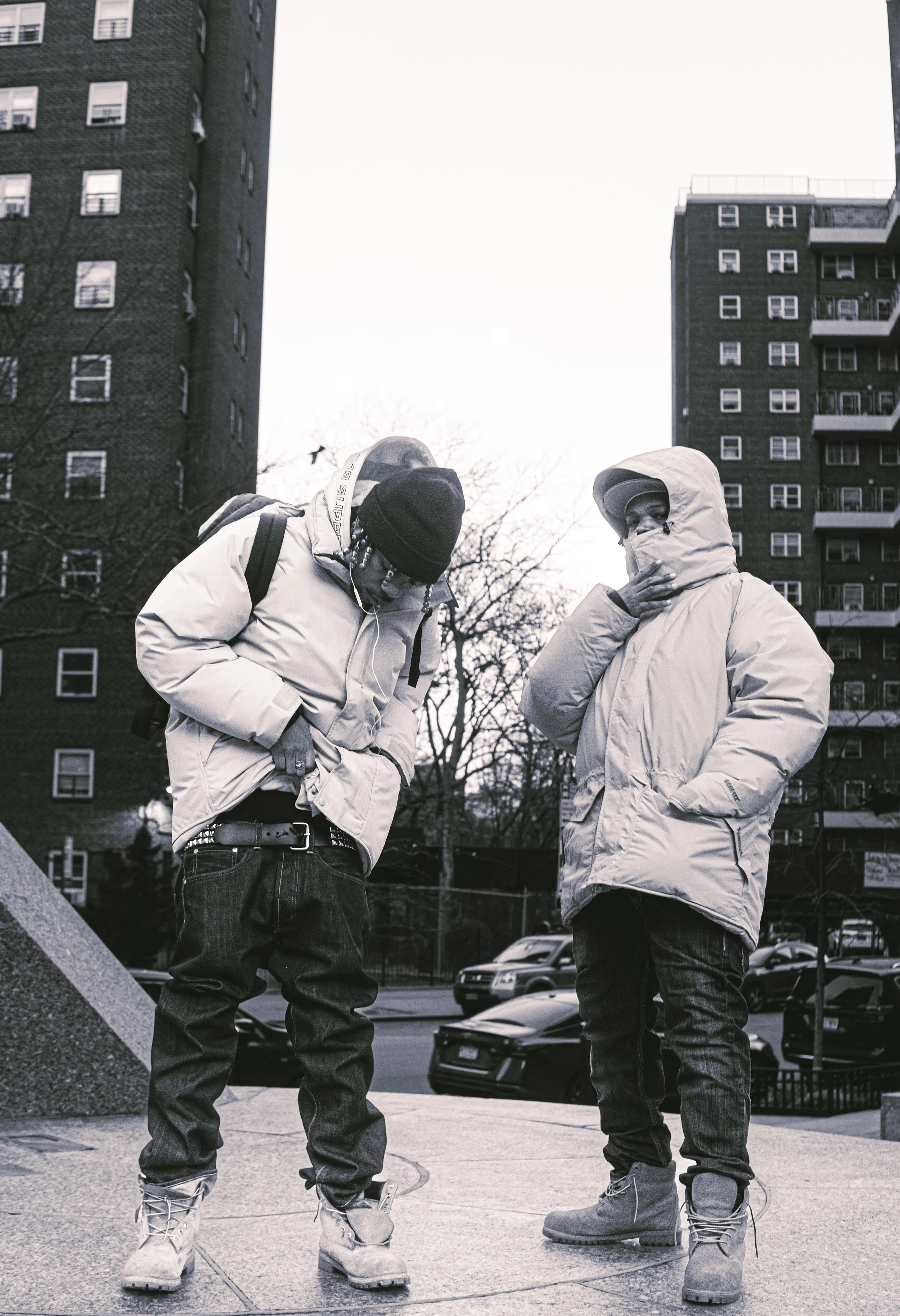 Two people wearing winter coats and boots standing outdoors on a city sidewalk, with tall apartment buildings and parked cars in the background.