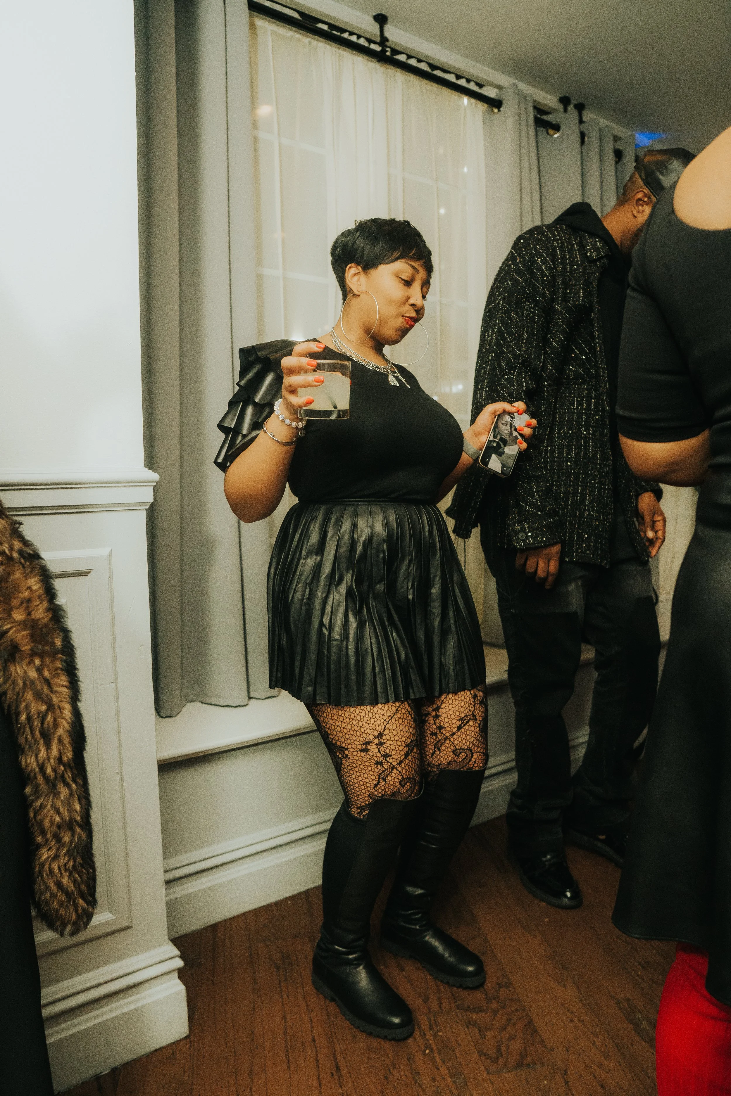 A woman holding a drink, wearing a black outfit with mesh stockings, and standing indoors.