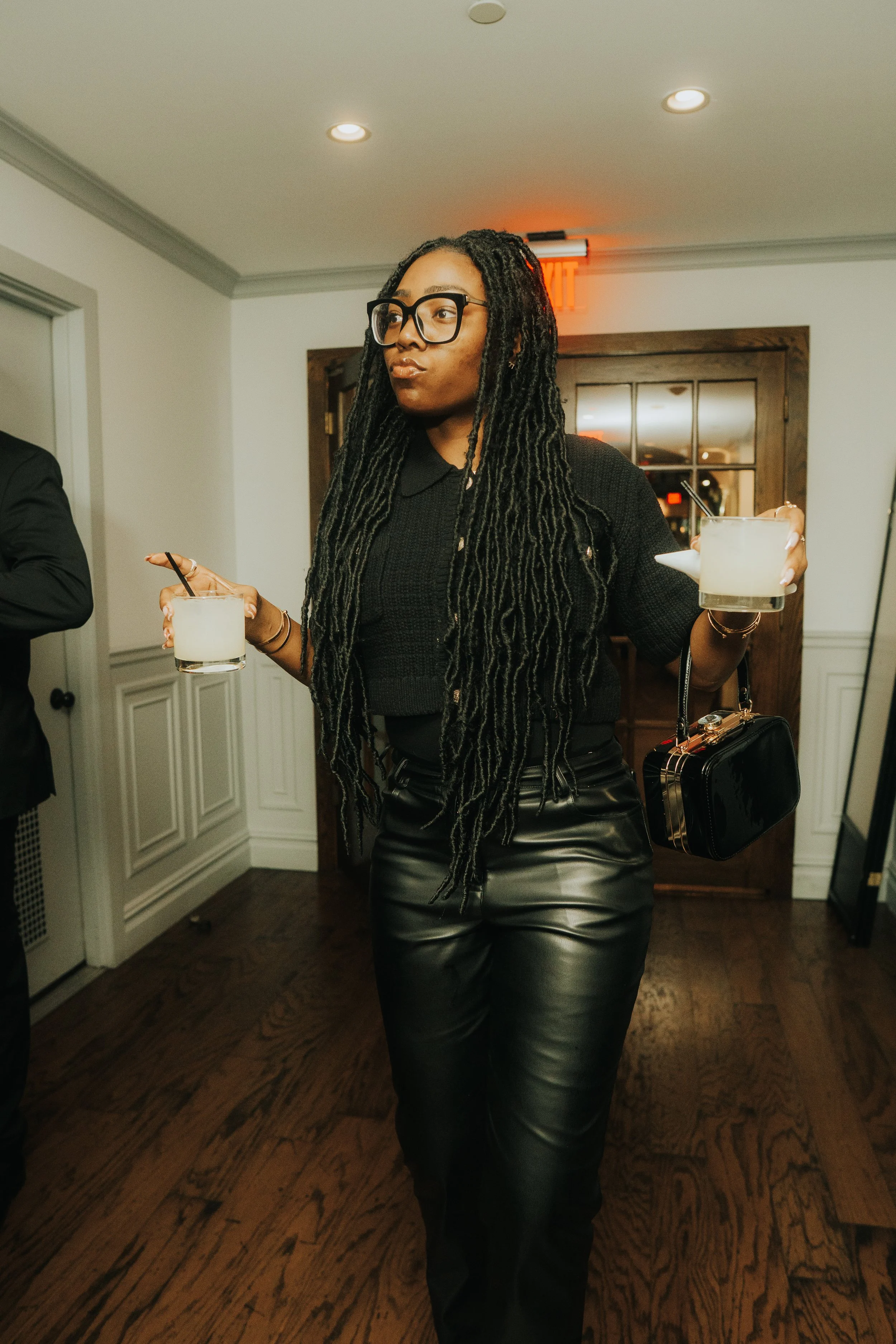 Person in black outfit holding two drinks at an indoor event.