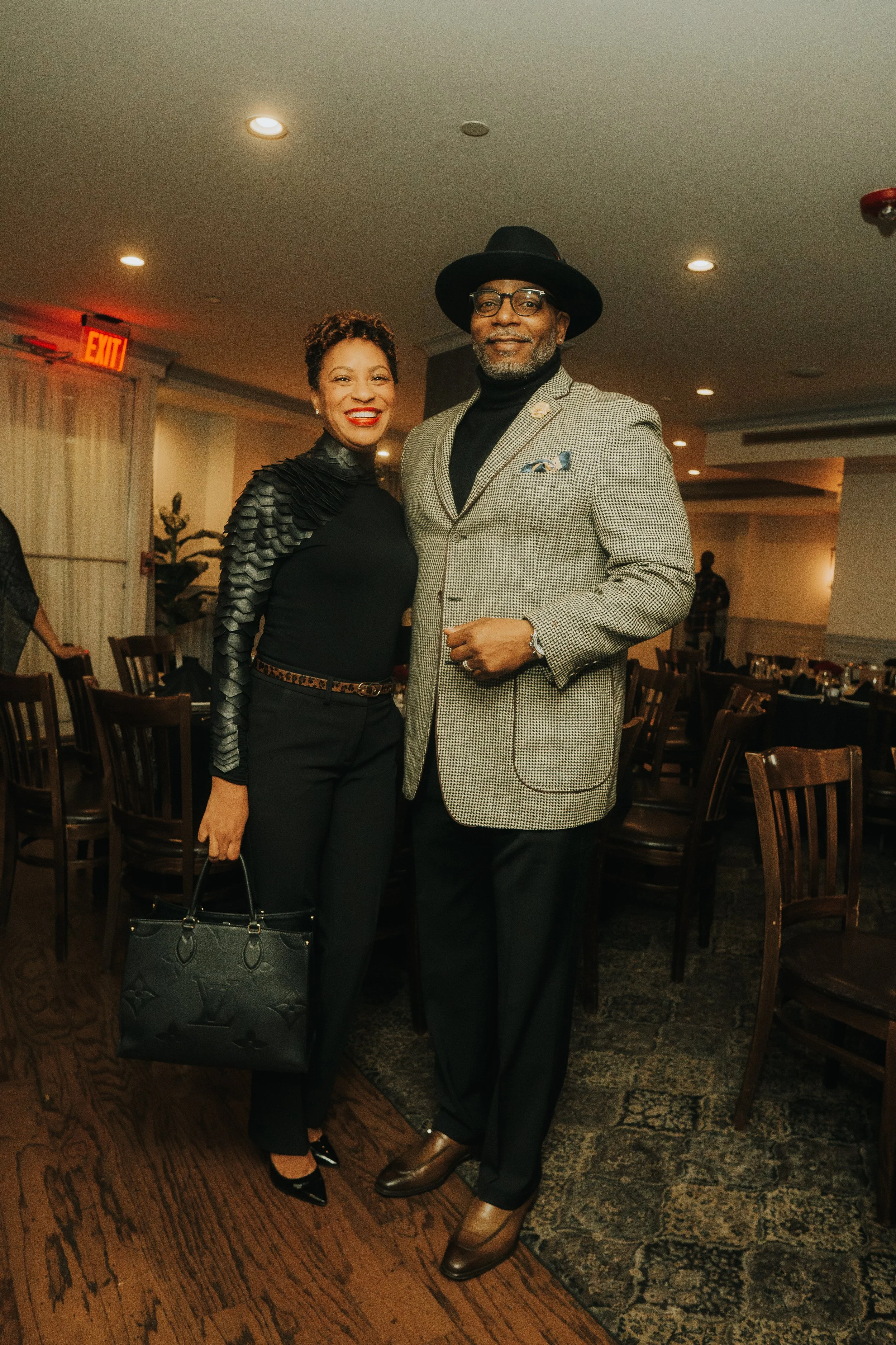 A man and woman posing in a dimly lit dining area, with chairs and tables in the background. The woman is wearing a stylish black outfit and holding a handbag. The man is wearing a gray checkered blazer, black hat, and glasses.