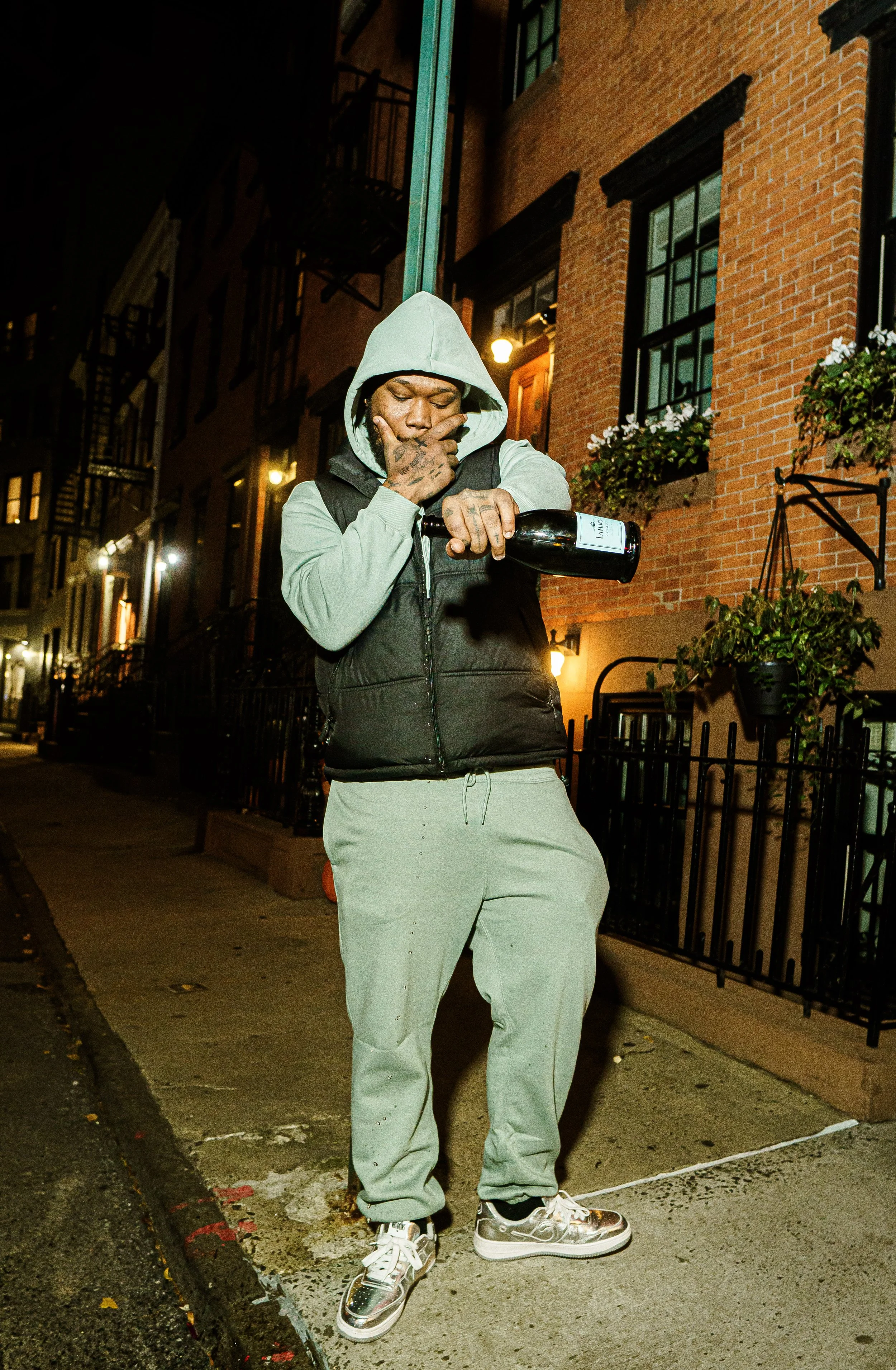 A man in a gray hoodie, black vest, and beige sweatpants stands on a city sidewalk at night, opening a bottle of wine, with an urban brick apartment building in the background.