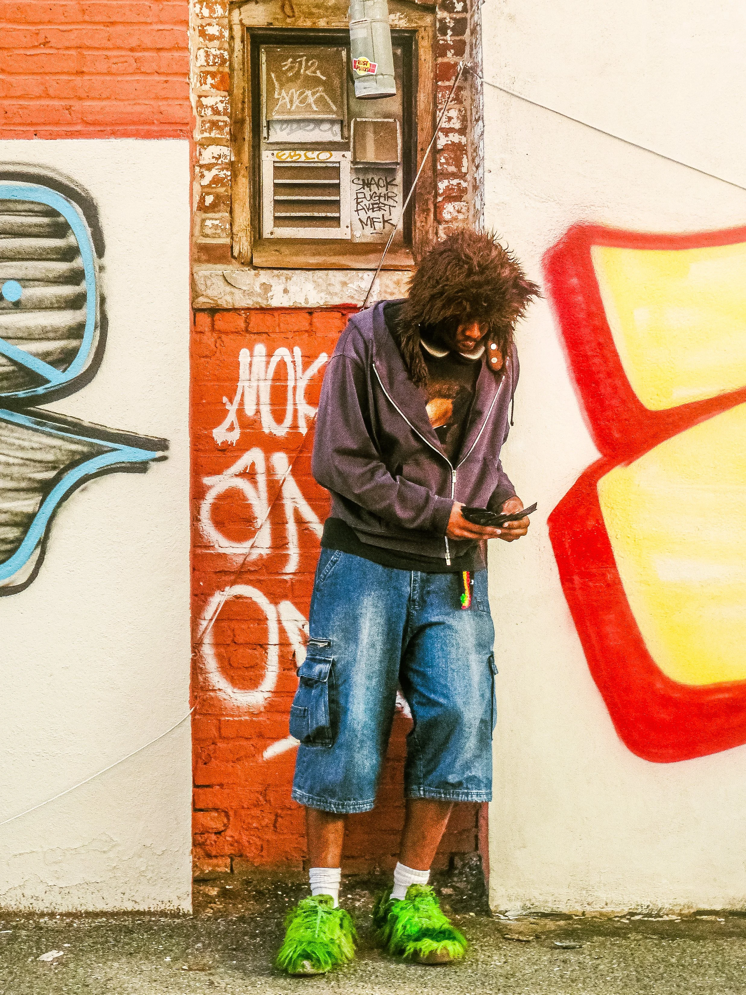 A young man with curly hair wearing bright green fuzzy shoes, white socks, baggy denim shorts, a dark hoodie, and a graphic t-shirt, standing in front of a graffiti-covered brick wall while looking at his phone.