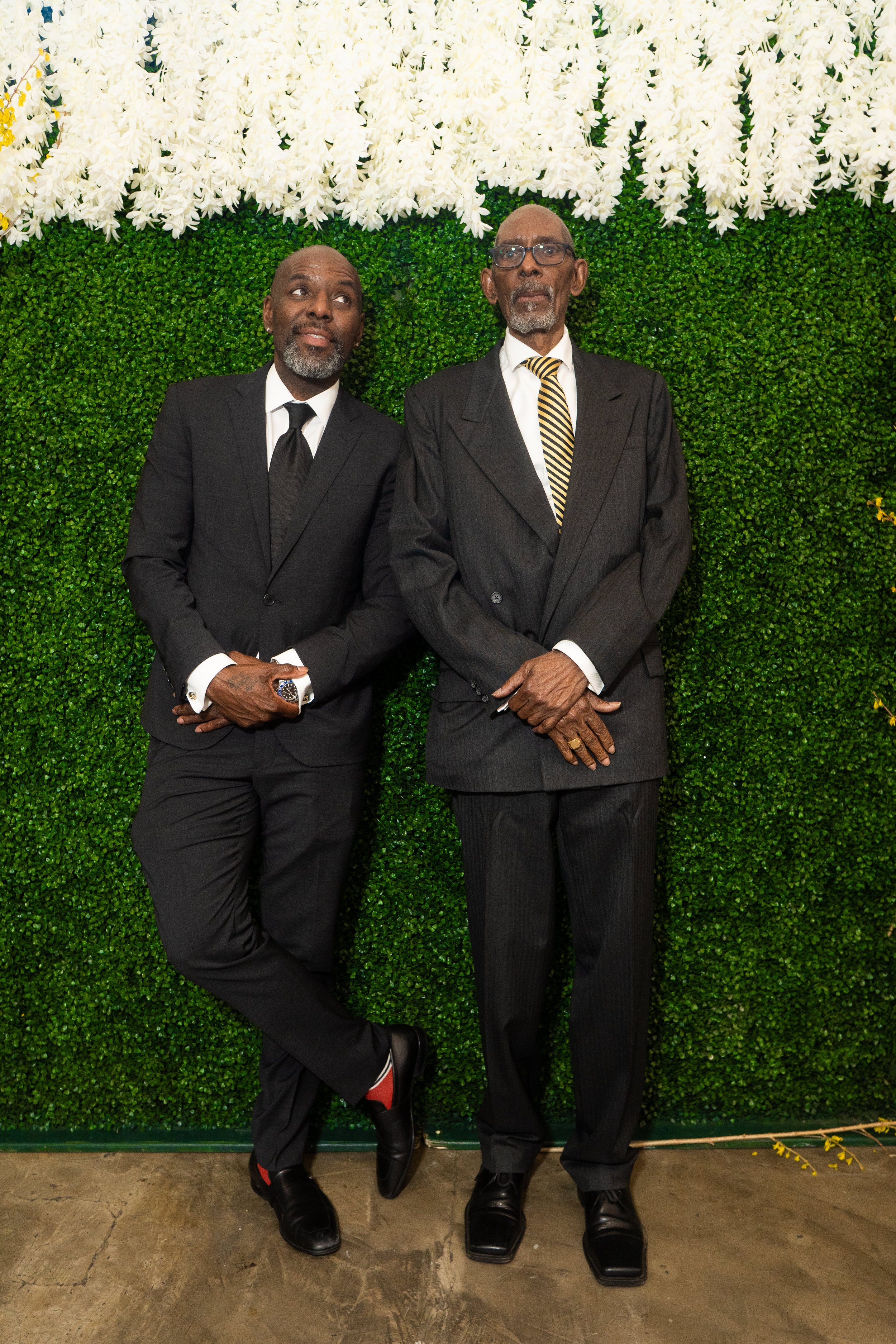 Two men in suits standing against a green ivy wall with white flowers.