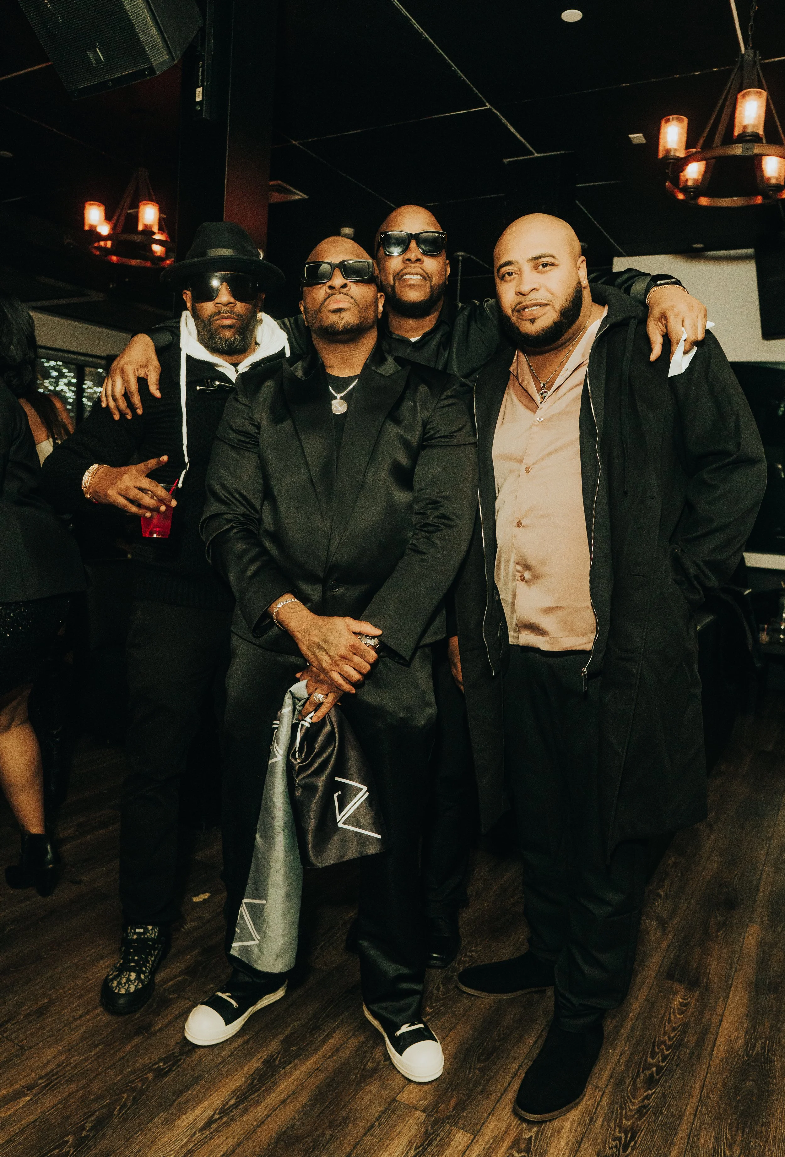 Four men posing together in a stylish, dimly-lit setting. They are wearing dark clothing and sunglasses, with one holding a drink. The atmosphere suggests a nightlife or celebration event.