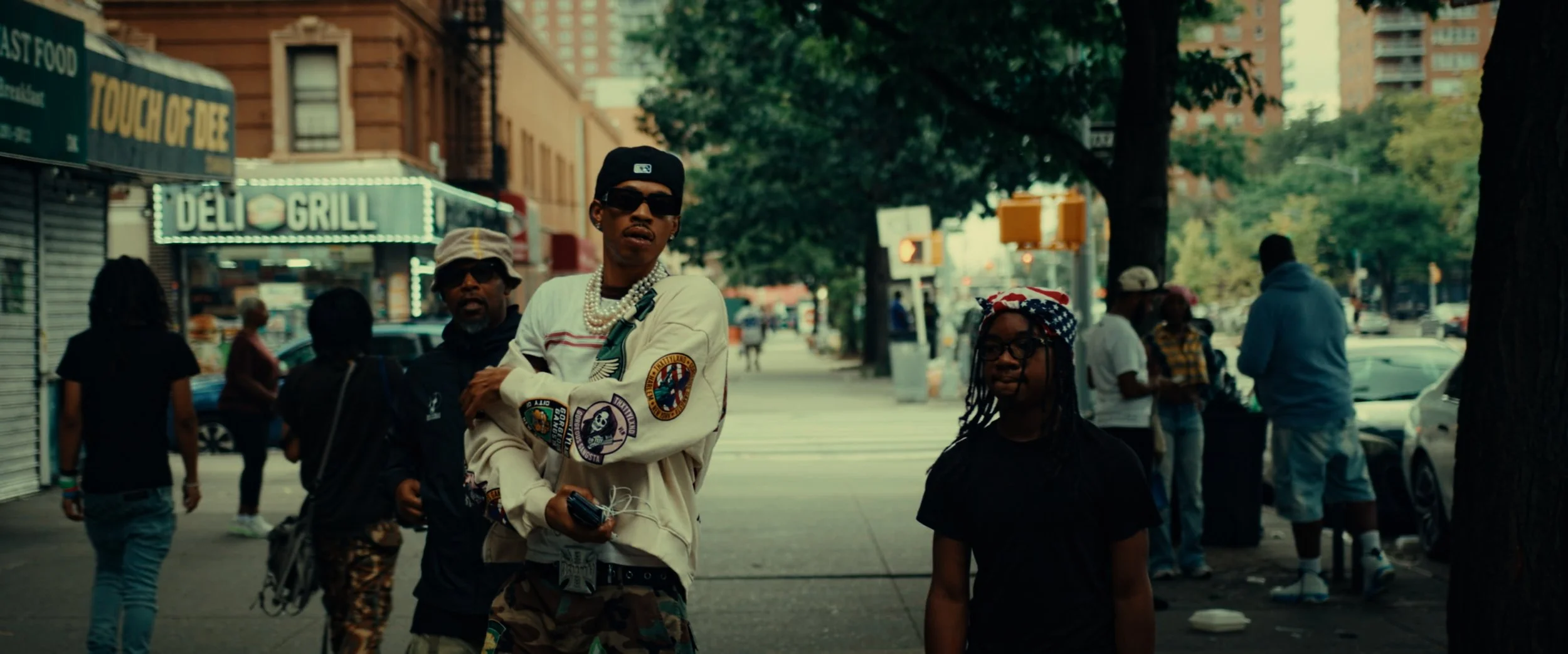 People standing on a city sidewalk, including a young man with sunglasses and layered chains, and a young girl with glasses and a red, white, and blue bandana, with storefronts and trees in the background.