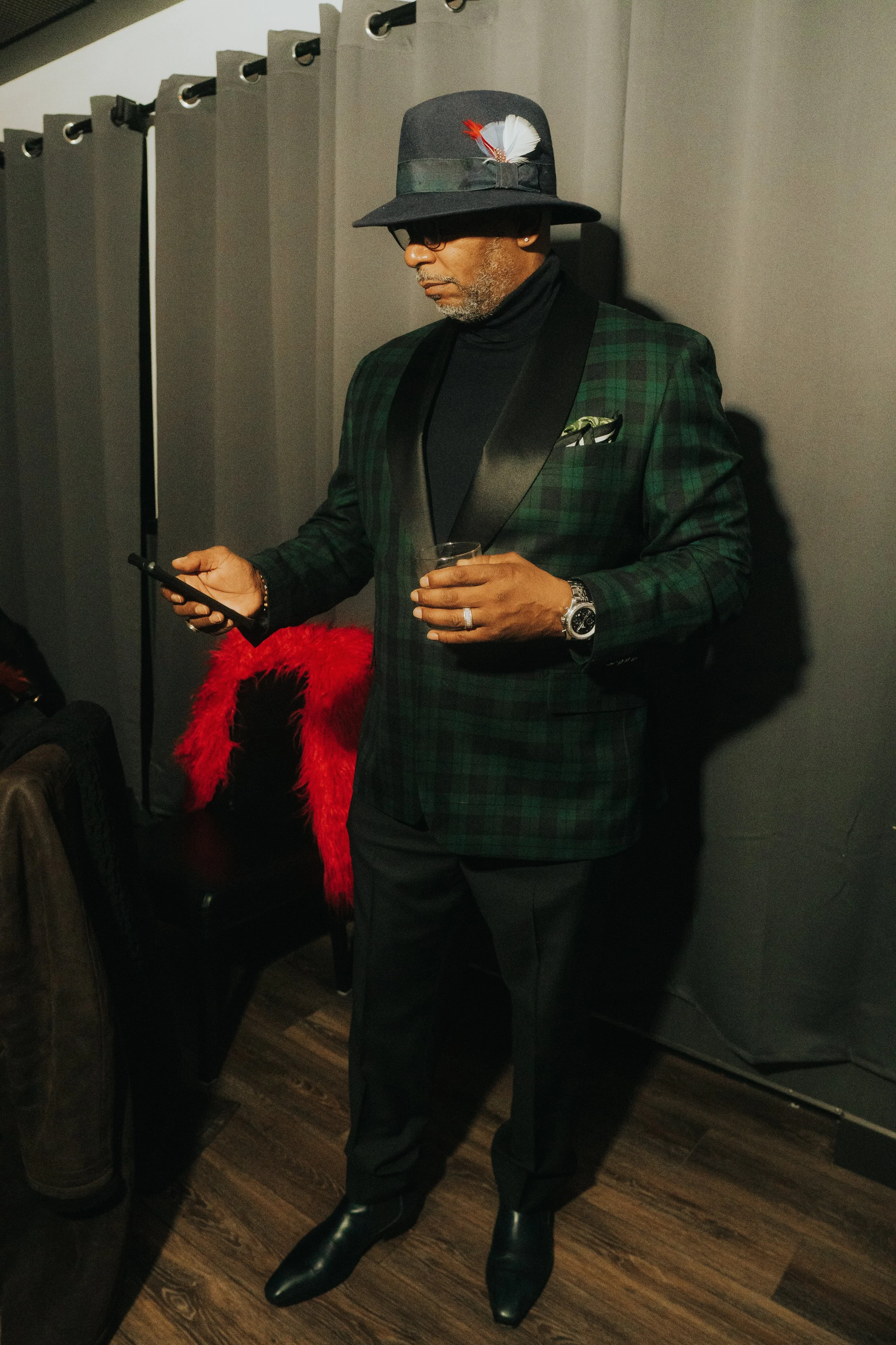 Man in plaid suit and hat, holding beverage and phone, standing indoors.