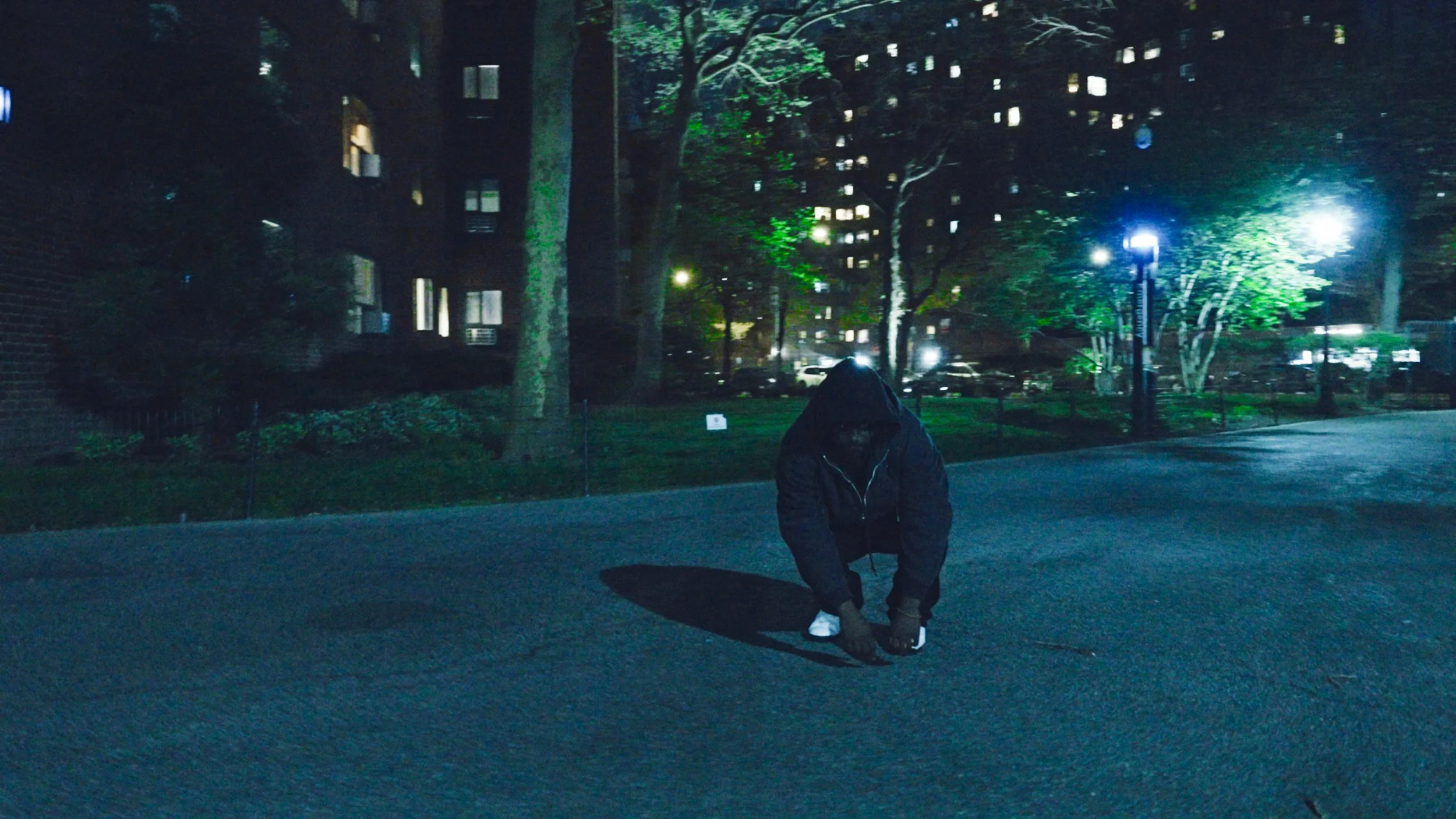 Person in a dark hoodie crouching on a city street at night, surrounded by buildings and trees illuminated by streetlights.