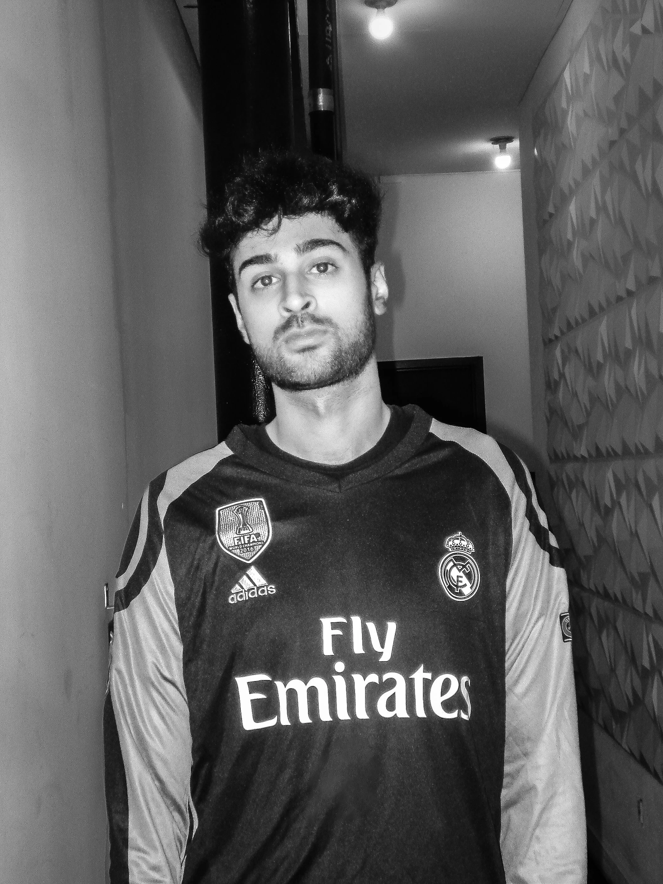 A young man with dark, curly hair and facial hair, wearing a Real Madrid soccer jersey in a black and white photo, standing indoors.