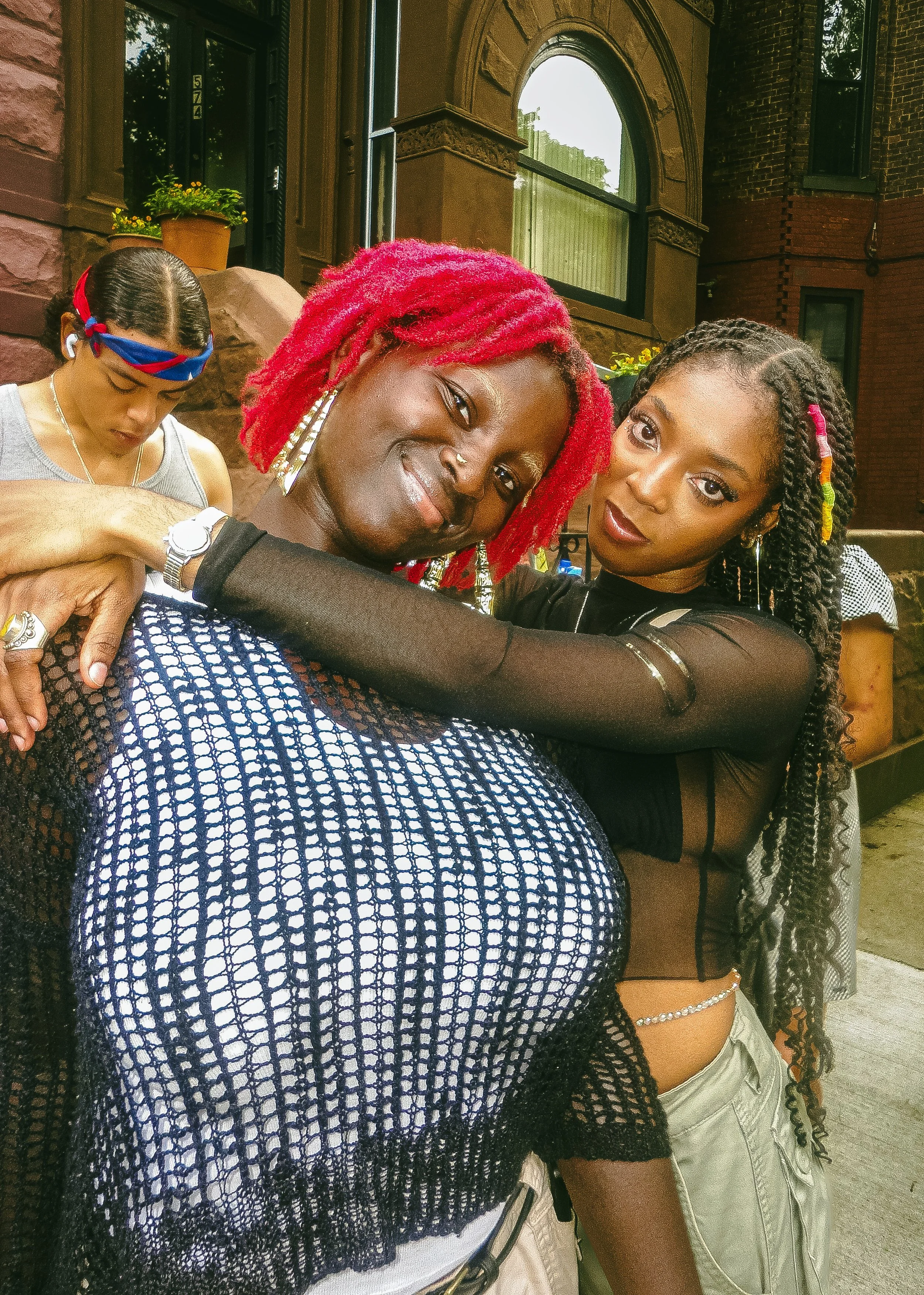 Two women hugging and smiling outdoors in front of a brownstone building, one with bright red dreadlocks and the other with long curly hair.