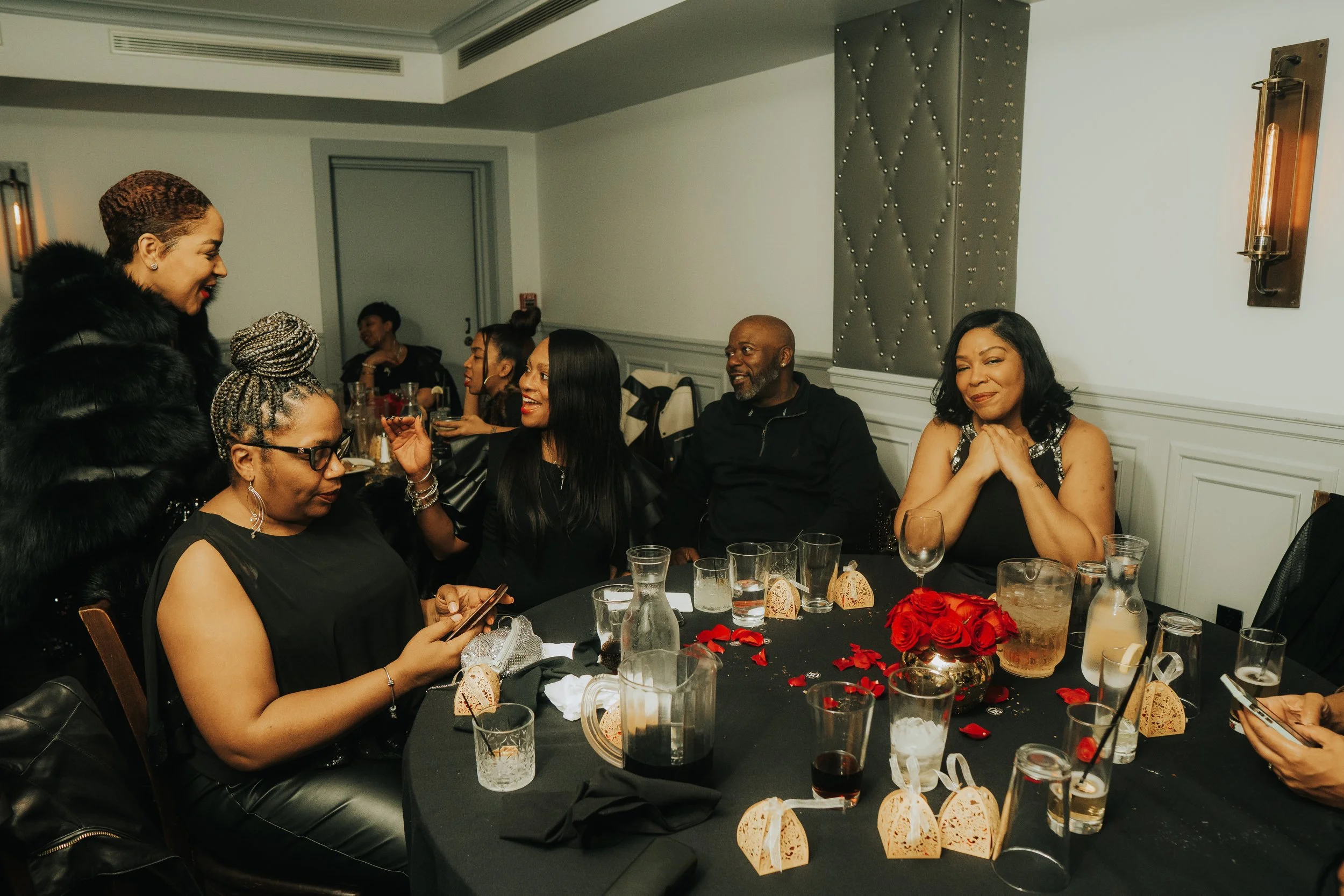 Group of people seated around a table, dressed in black, at an indoor event. The table is decorated with glasses, small gift boxes, rose petals, and a centerpiece of red roses. One person is using a smartphone.