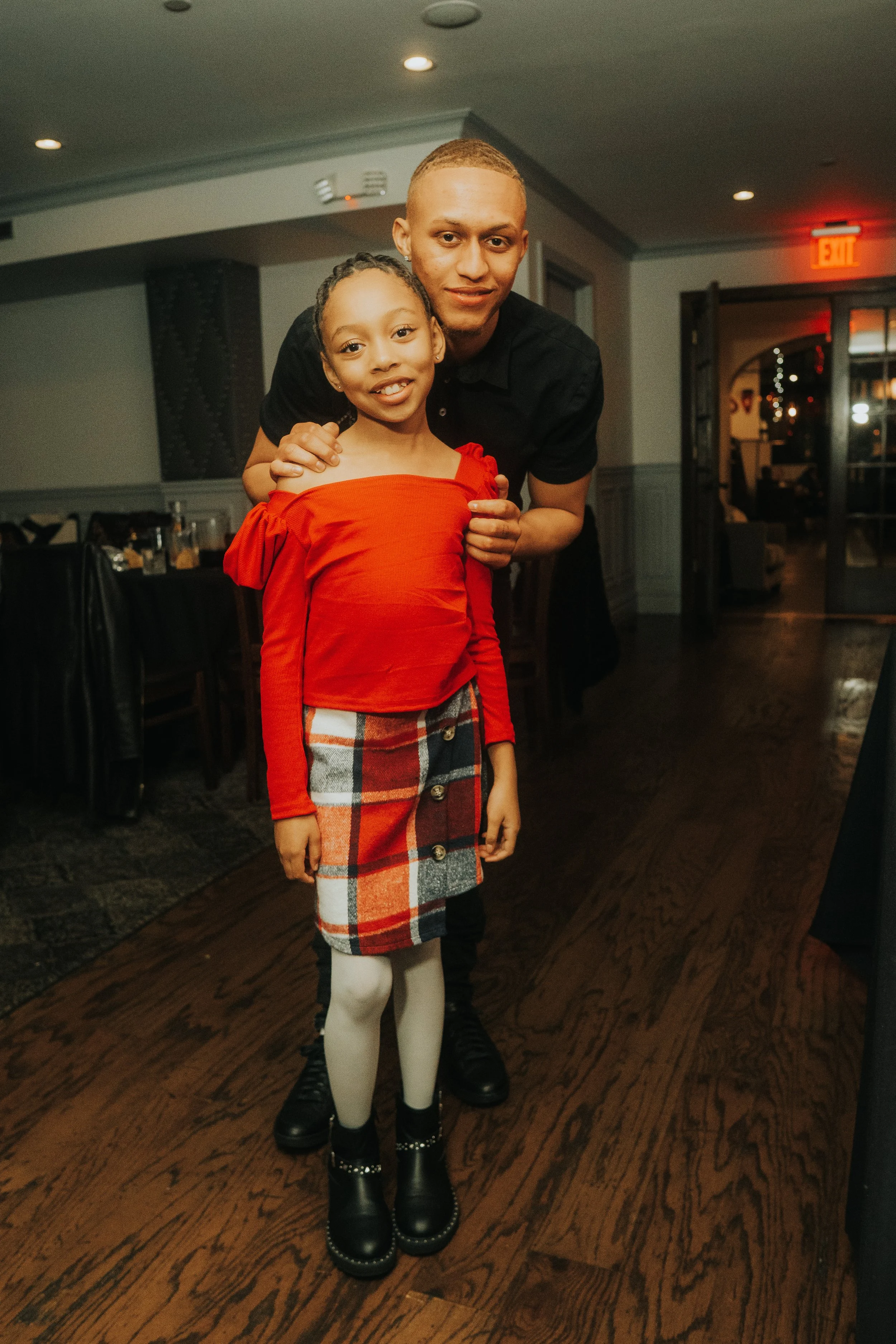 A young girl in a red top and plaid skirt poses with an older individual in a black shirt inside a warmly lit room.