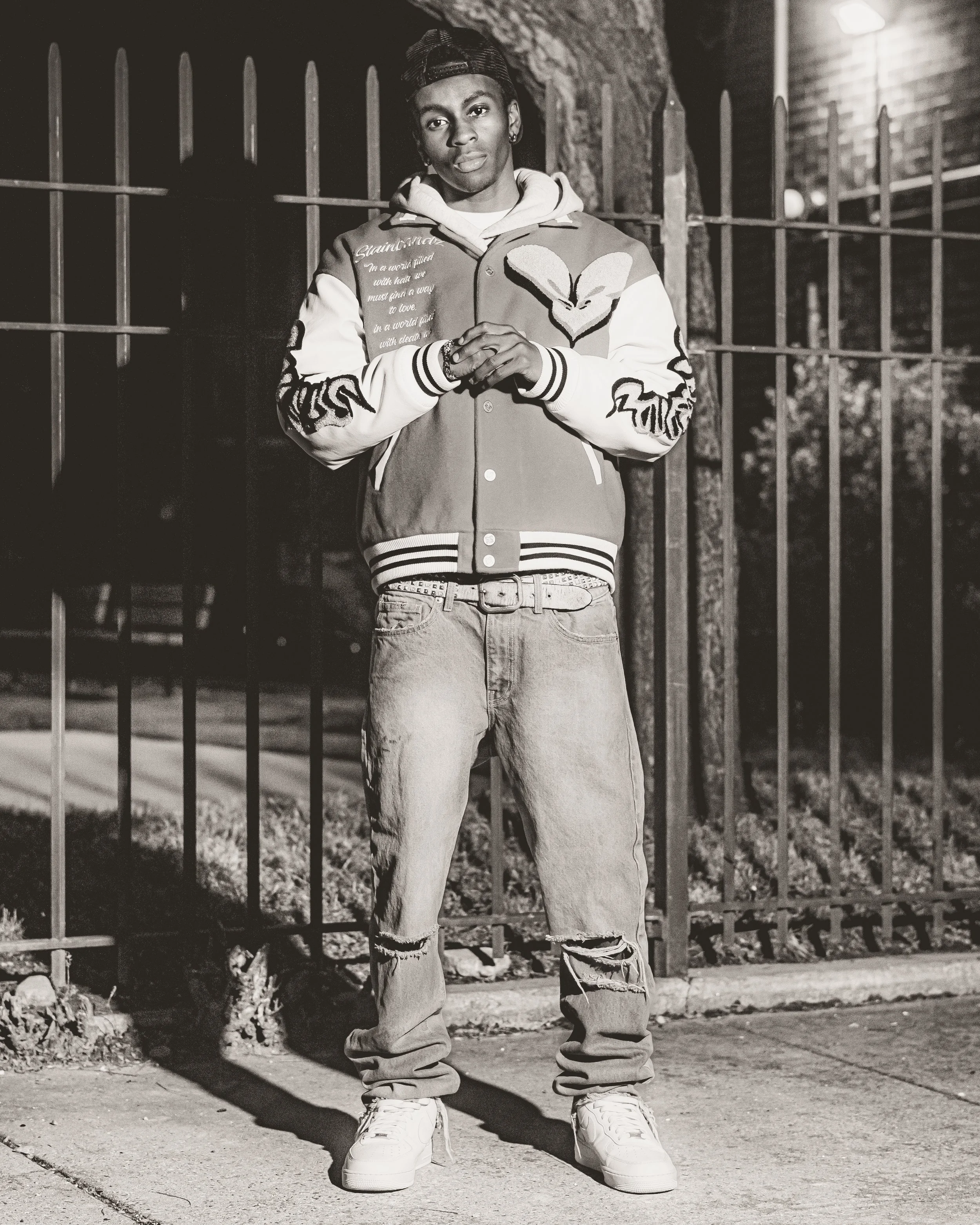 A young man standing outdoors at night beside a metal fence, wearing a varsity jacket, ripped jeans, sneakers, a backwards cap, with a serious expression.