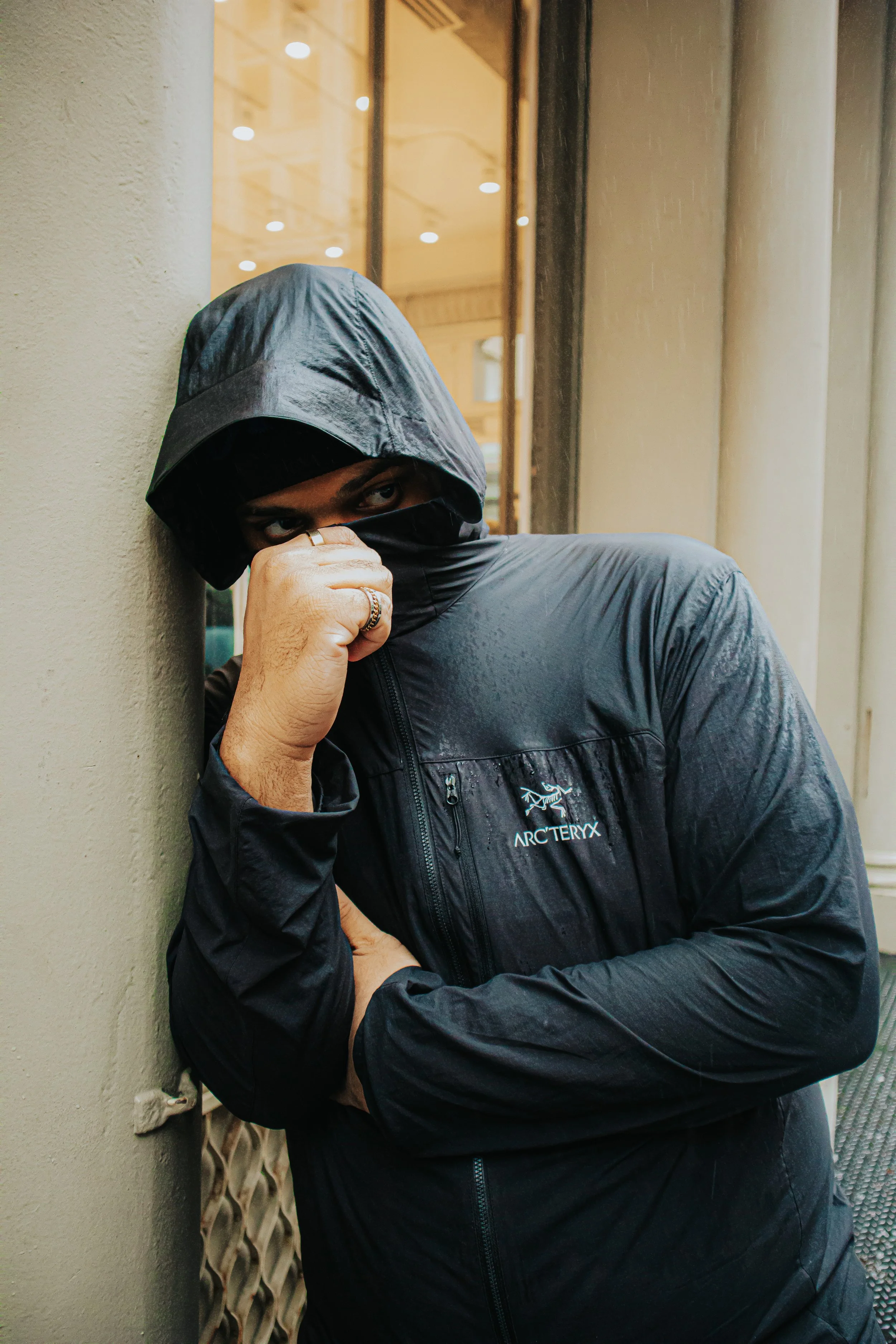 A person wearing a black Arc'teryx jacket with the hood up, standing against a wall, partially covering their face with the jacket, looking to the side.