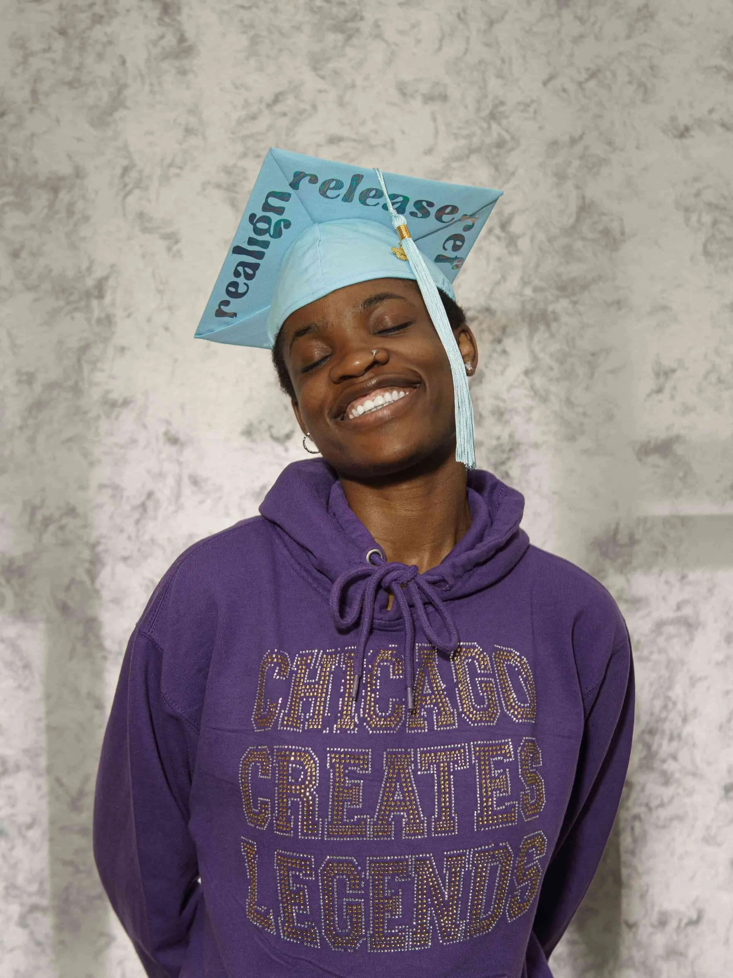 A smiling woman with short hair, earrings, and a nose ring wearing a purple hoodie with the words 'CHICAGO GREATES LEGENDS' in rhinestones, standing with eyes closed against a gray marbled background. She wears a light blue graduation cap with words 'released' and 'real' written on it.