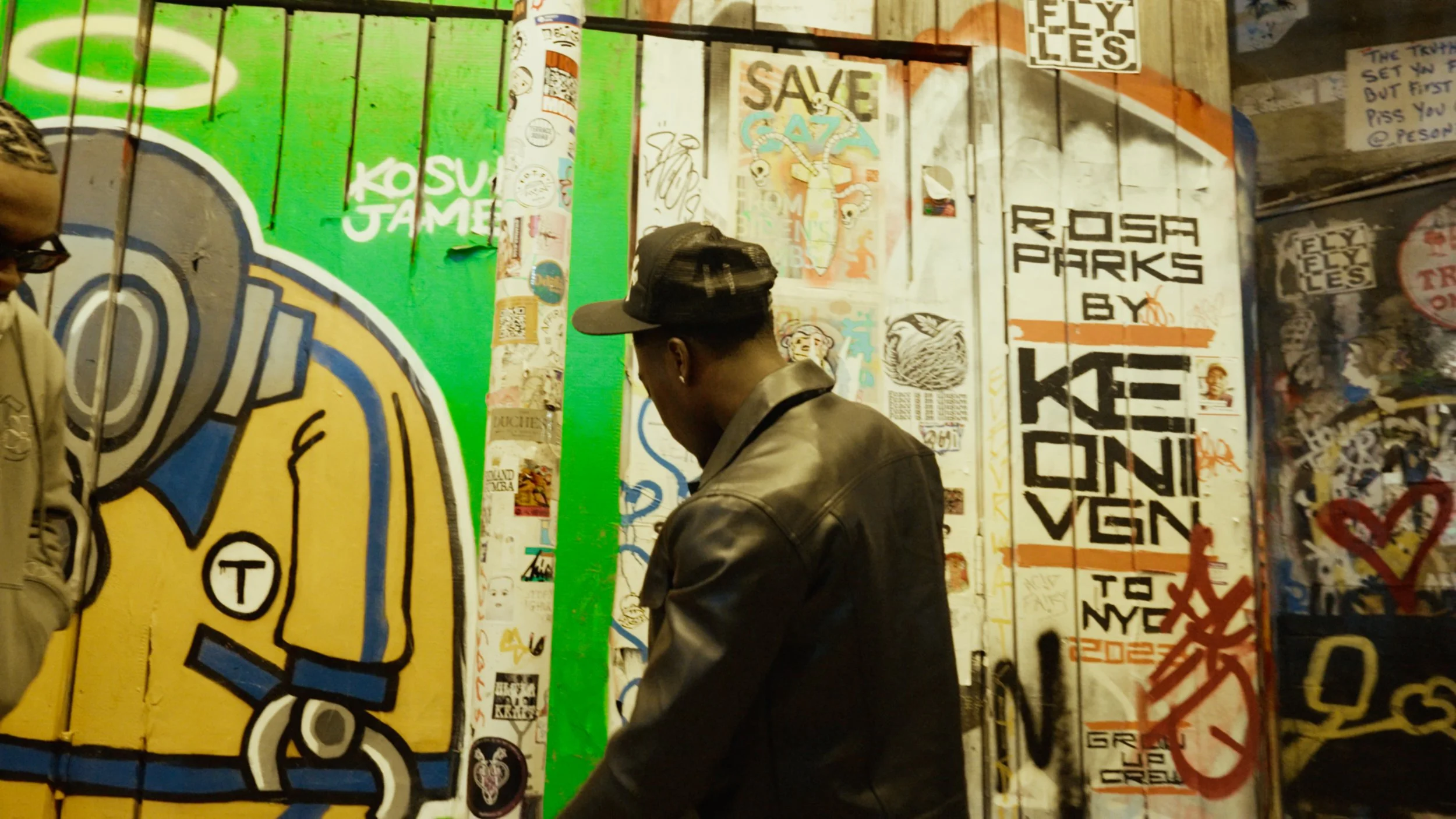 Two people standing by a wall covered in colorful street art and graffiti, featuring a large astronaut mural and various stickers and posters.