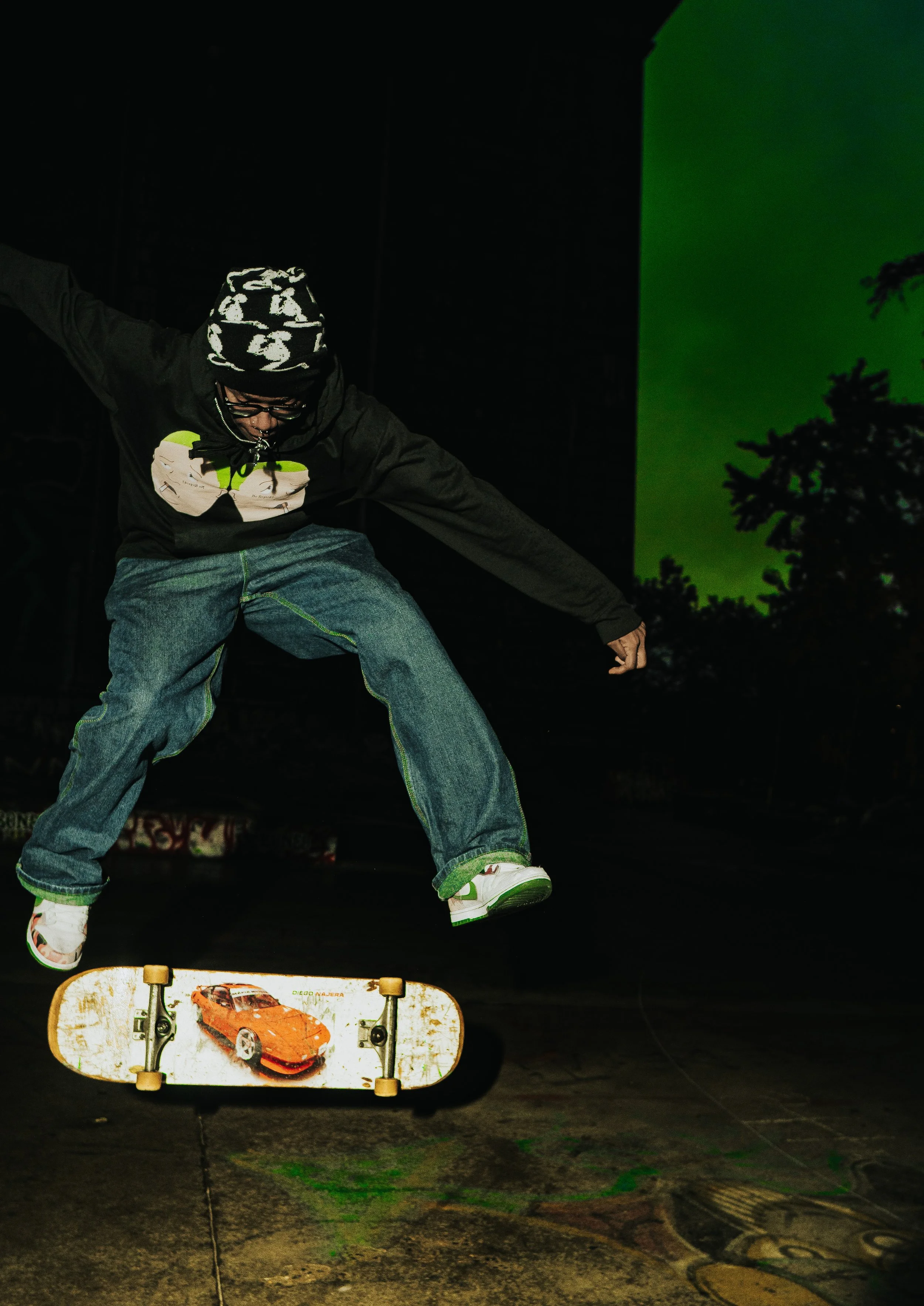 A person skateboarding at night, wearing a black hoodie, jeans, and a beanie with a white camouflage pattern, on a skateboard with a red sports car graphic, with a green background projected on a wall.