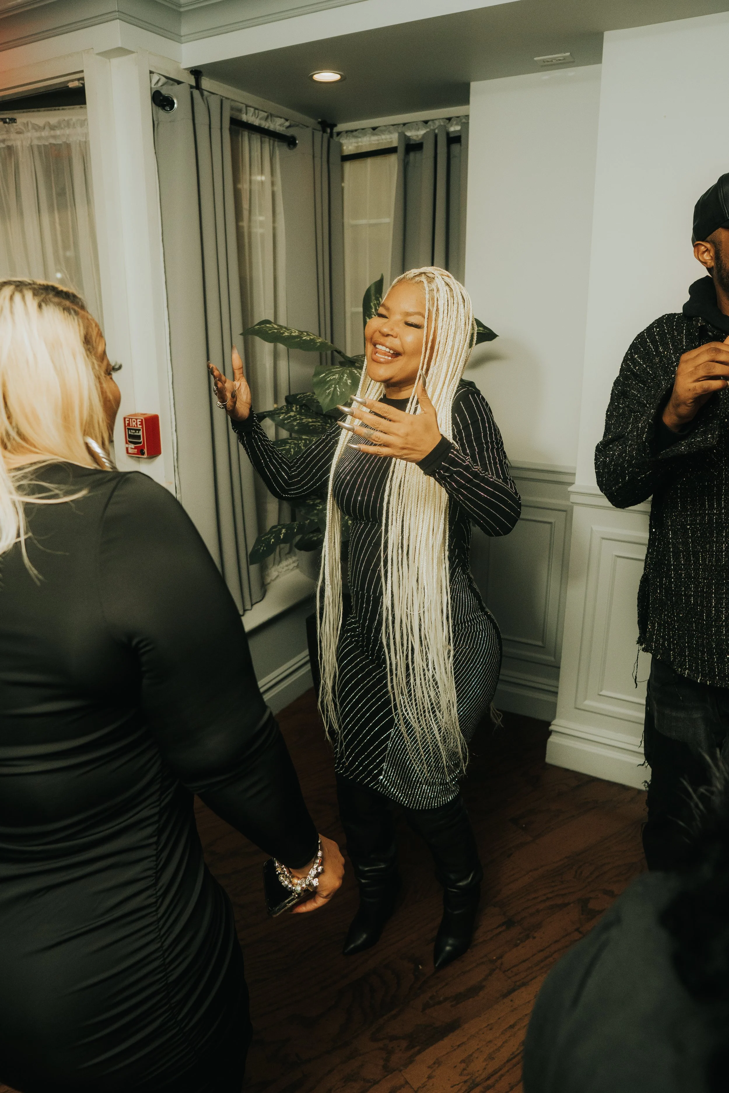 A group of people engaged in conversation in a room with wooden flooring and curtains. One person with long blonde braids is smiling and gesturing, wearing a black outfit with silver accents. Another person stands nearby, partially visible.