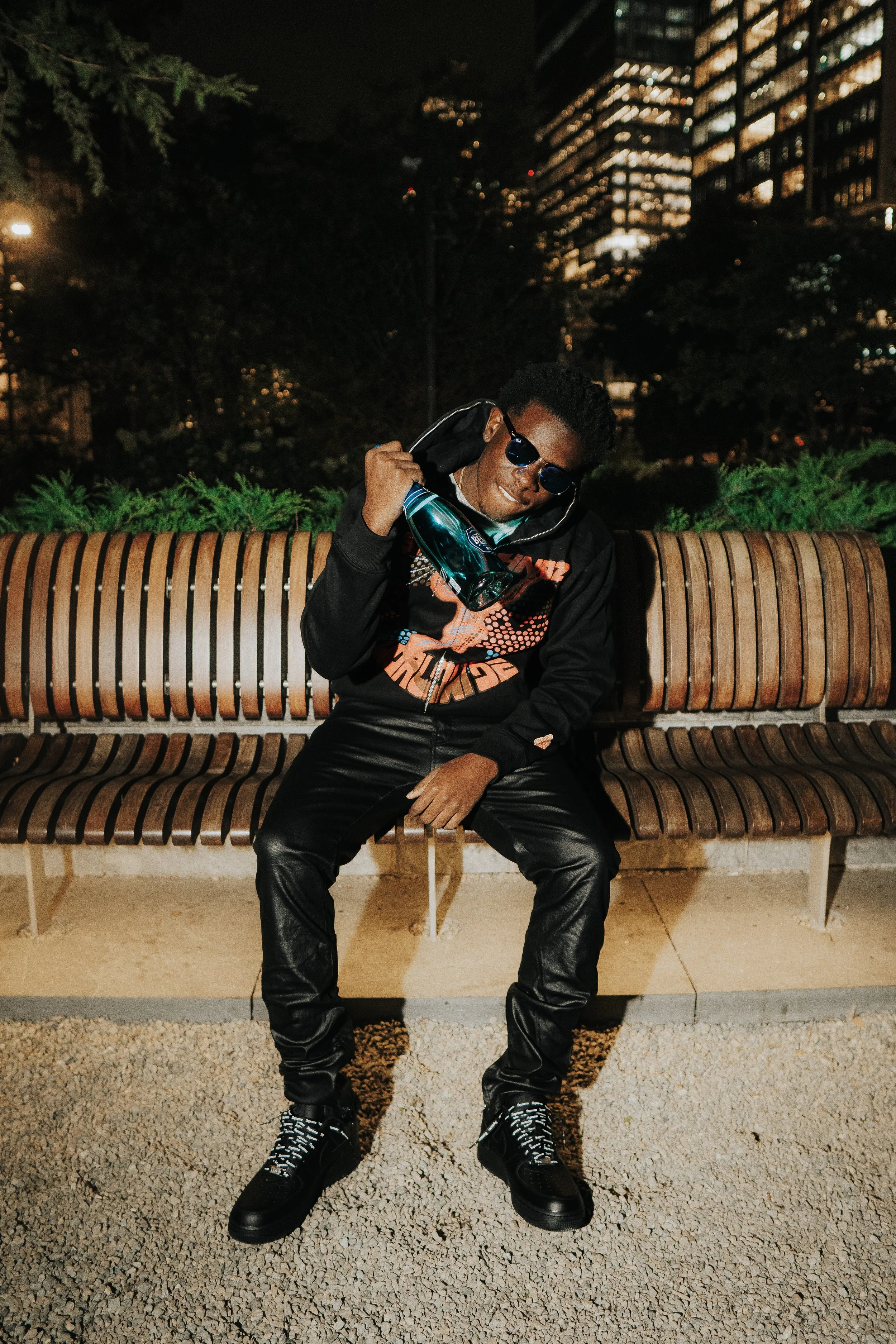 A person sitting on a wooden park bench at night, wearing sunglasses, black leather pants, boots, and holding a bottle of blue liquid. The background features city buildings illuminated at night.