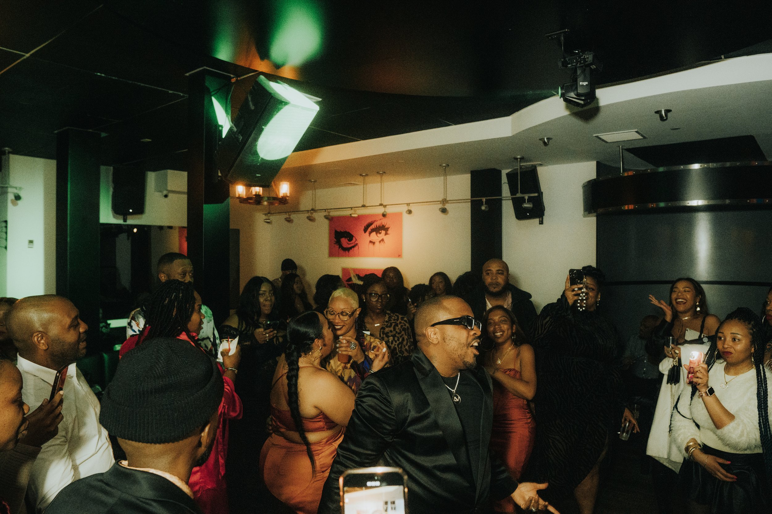 A lively indoor party with a diverse group of people dancing and taking photos, dressed in party attire. The venue has ambient green lighting and a framed artwork of eyes on the wall.