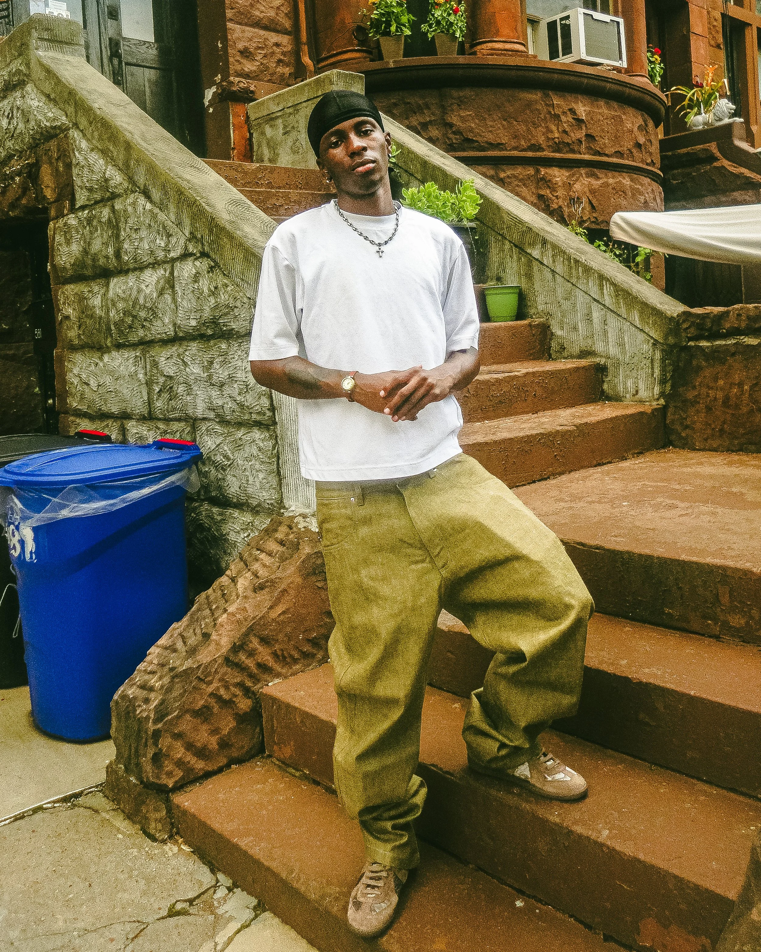 Young man standing on outdoor stairs wearing a black durag, white t-shirt, green baggy pants, and sneakers, with a chain necklace and a watch, in front of a stone wall and potted plants.