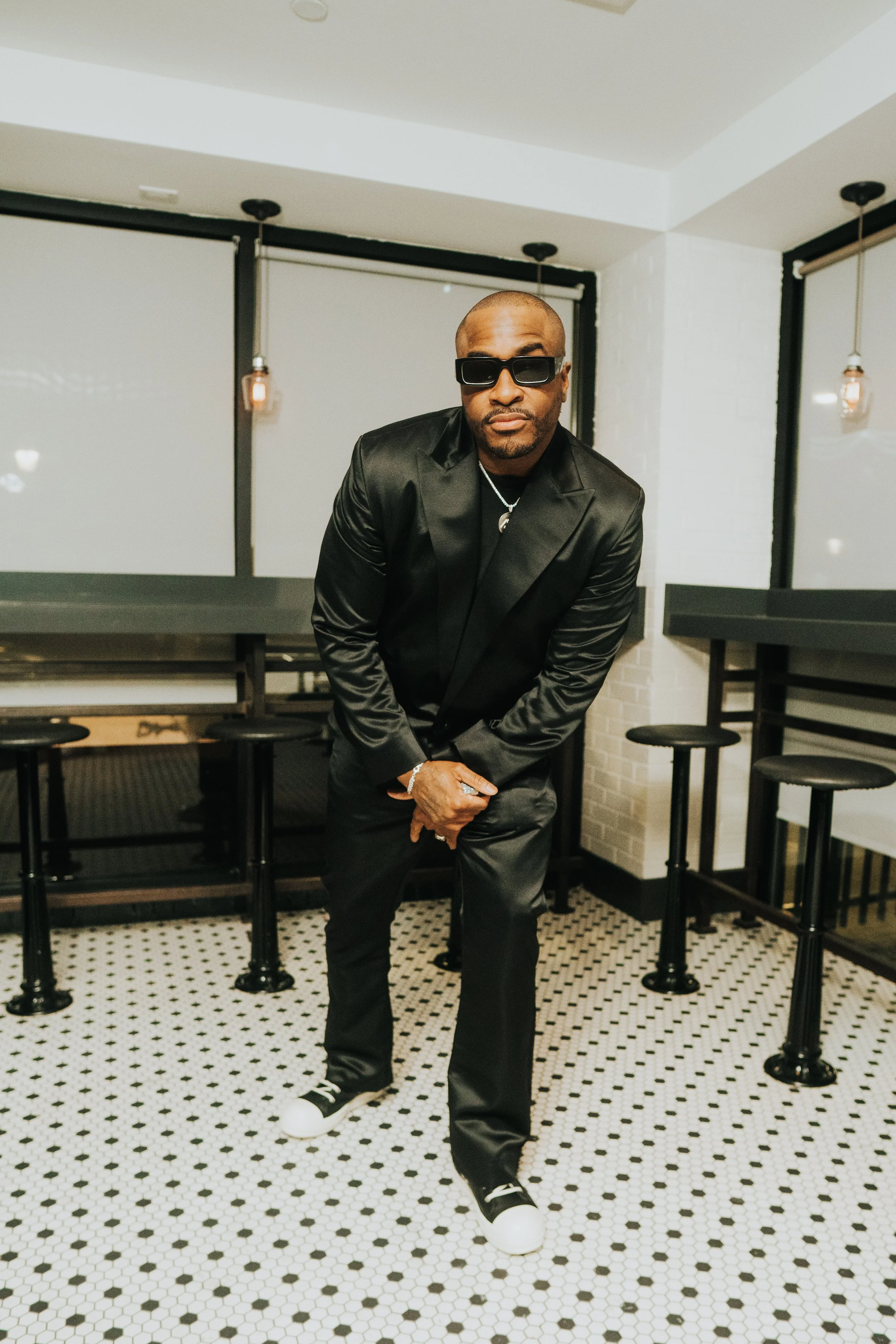 Man in black suit and sunglasses posing indoors on a black and white tiled floor.
