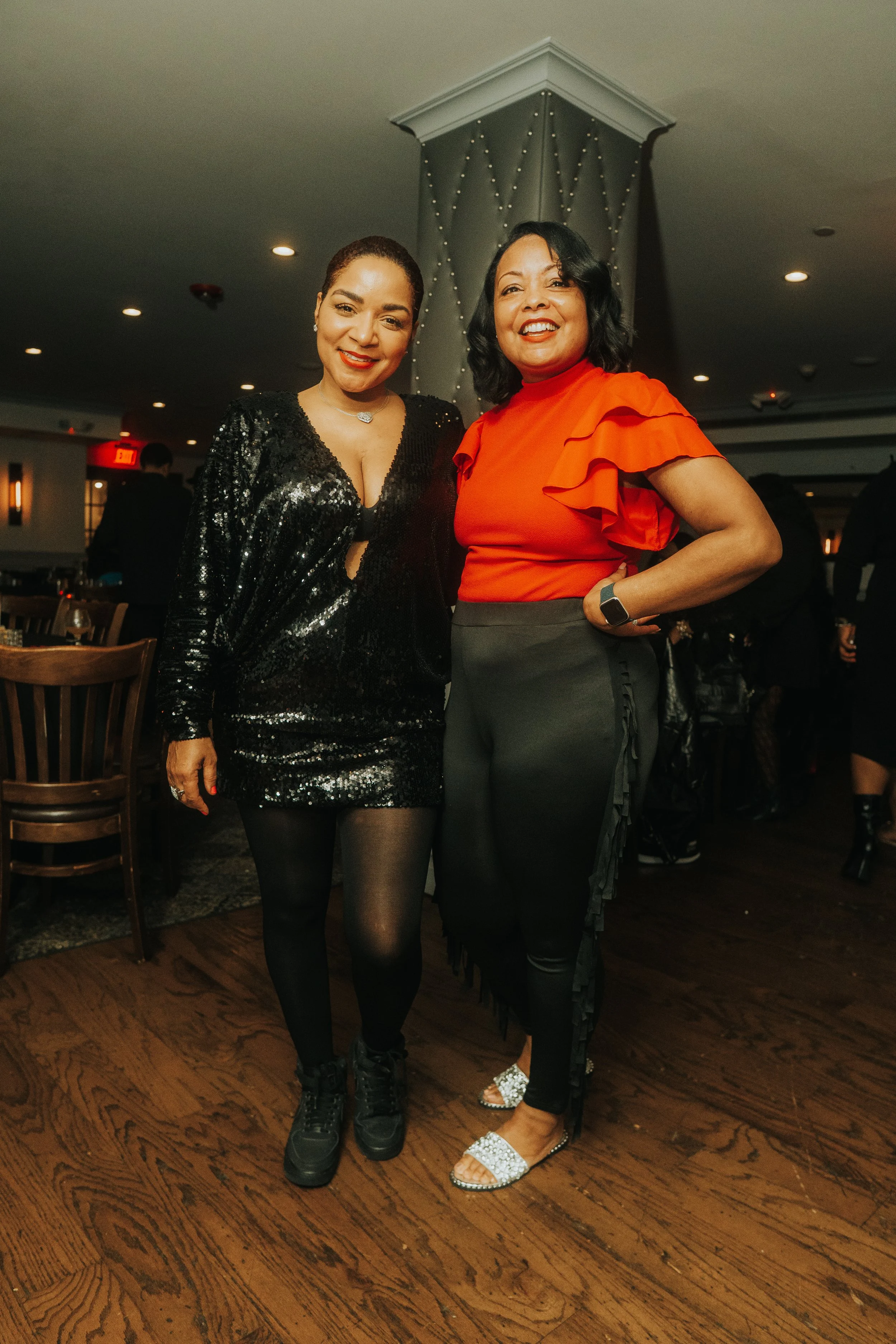 Two women posing together indoors at a social event. One wears a black sequin dress, the other a red blouse with black pants. Both are smiling and standing on wooden flooring.
