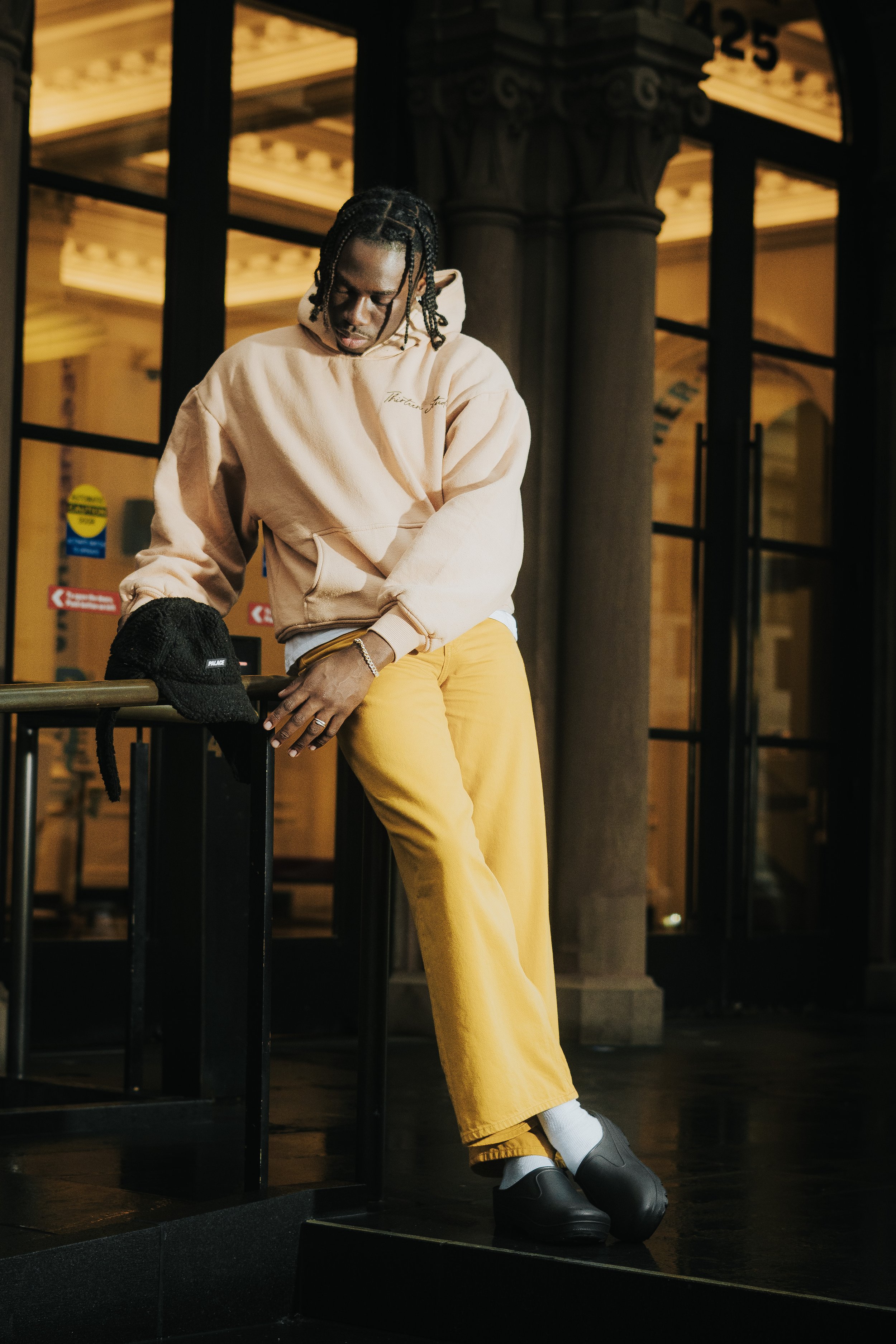 A person with braided hair standing indoors at night, wearing a beige hoodie, yellow pants, and black shoes, with a black beanie on the table, in front of a glass door with black frames and decorative columns.