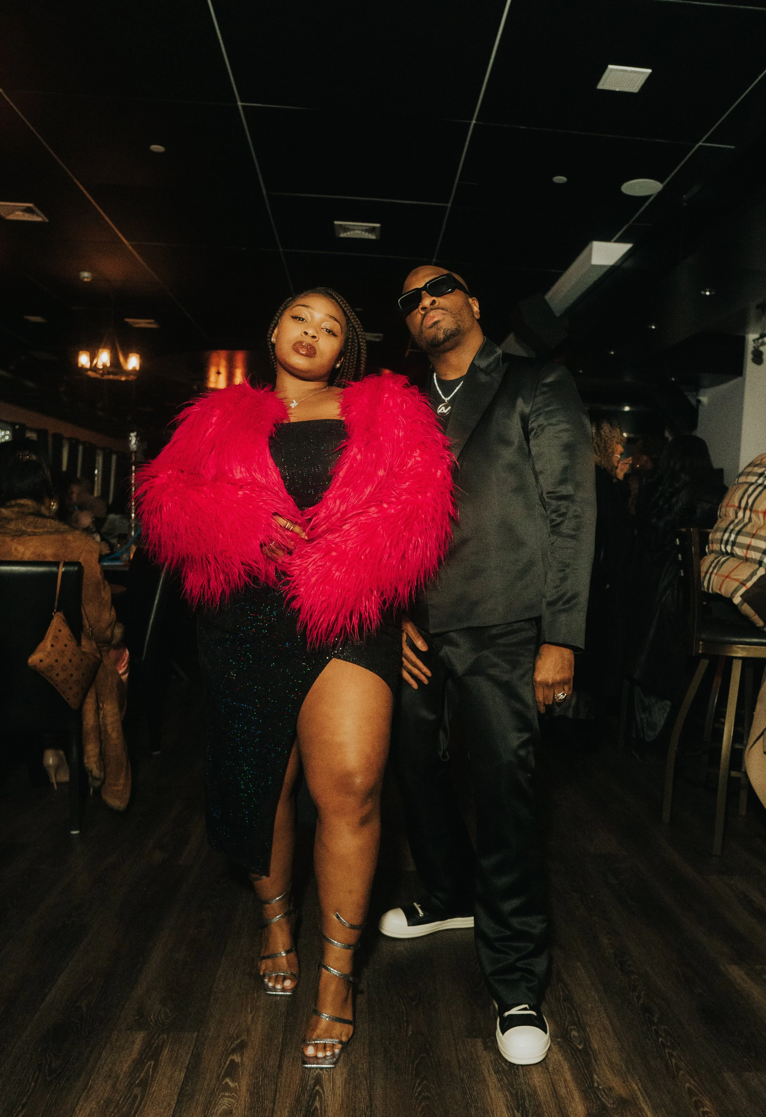 Two stylishly dressed people posing confidently indoors; one in a bright red feathered jacket and black dress with strappy heels, the other in a dark suit with sunglasses.