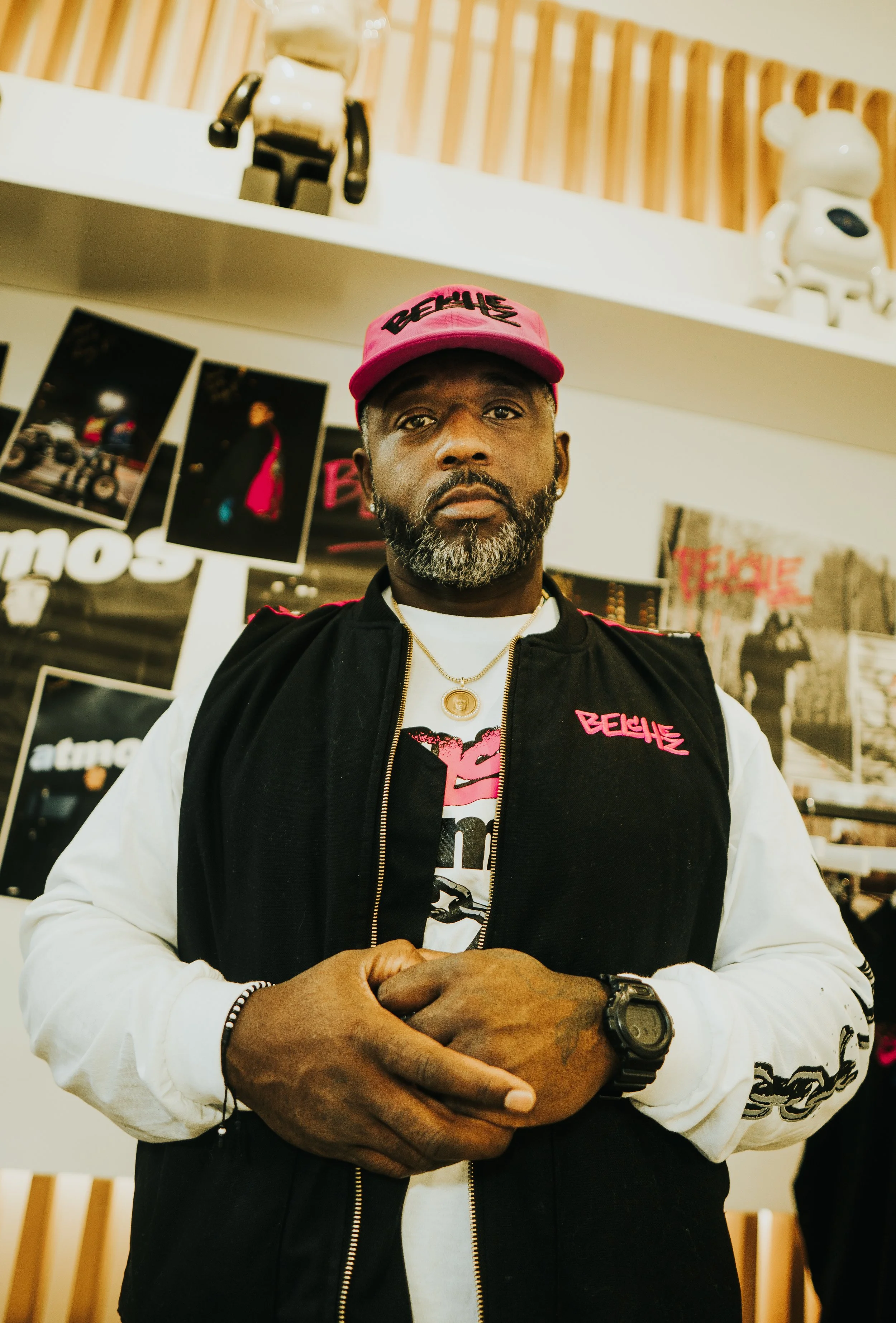 A man with a gray beard wearing a pink cap and a black and white hoodie with the word "Beyoncé" on it, standing in front of a display with photos and merchandise.