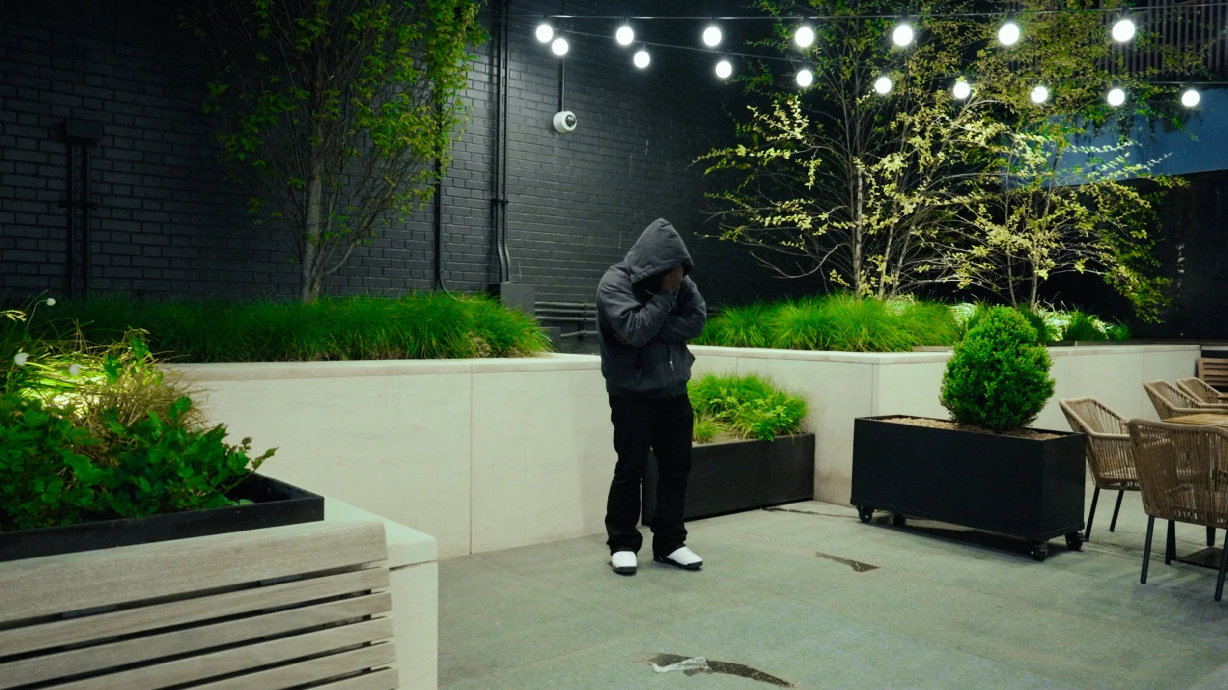A person in a dark hoodie standing in an outdoor area with string lights above, surrounded by modern planters holding green plants and bushes. The ground is paved and there are some outdoor chairs to the right.