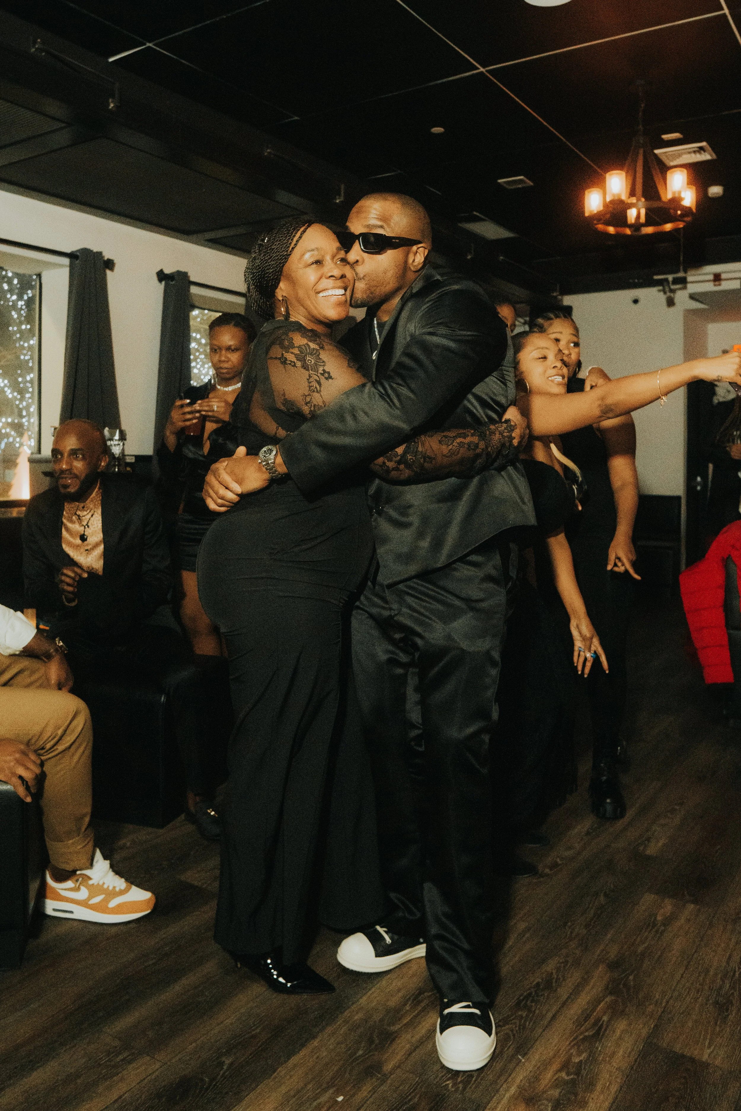 A lively indoor party scene with people dressed in formal black attire. A man wearing sunglasses kisses a smiling woman as they embrace in the center of the room. Other guests are visible in the background, some holding drinks and others taking photo
