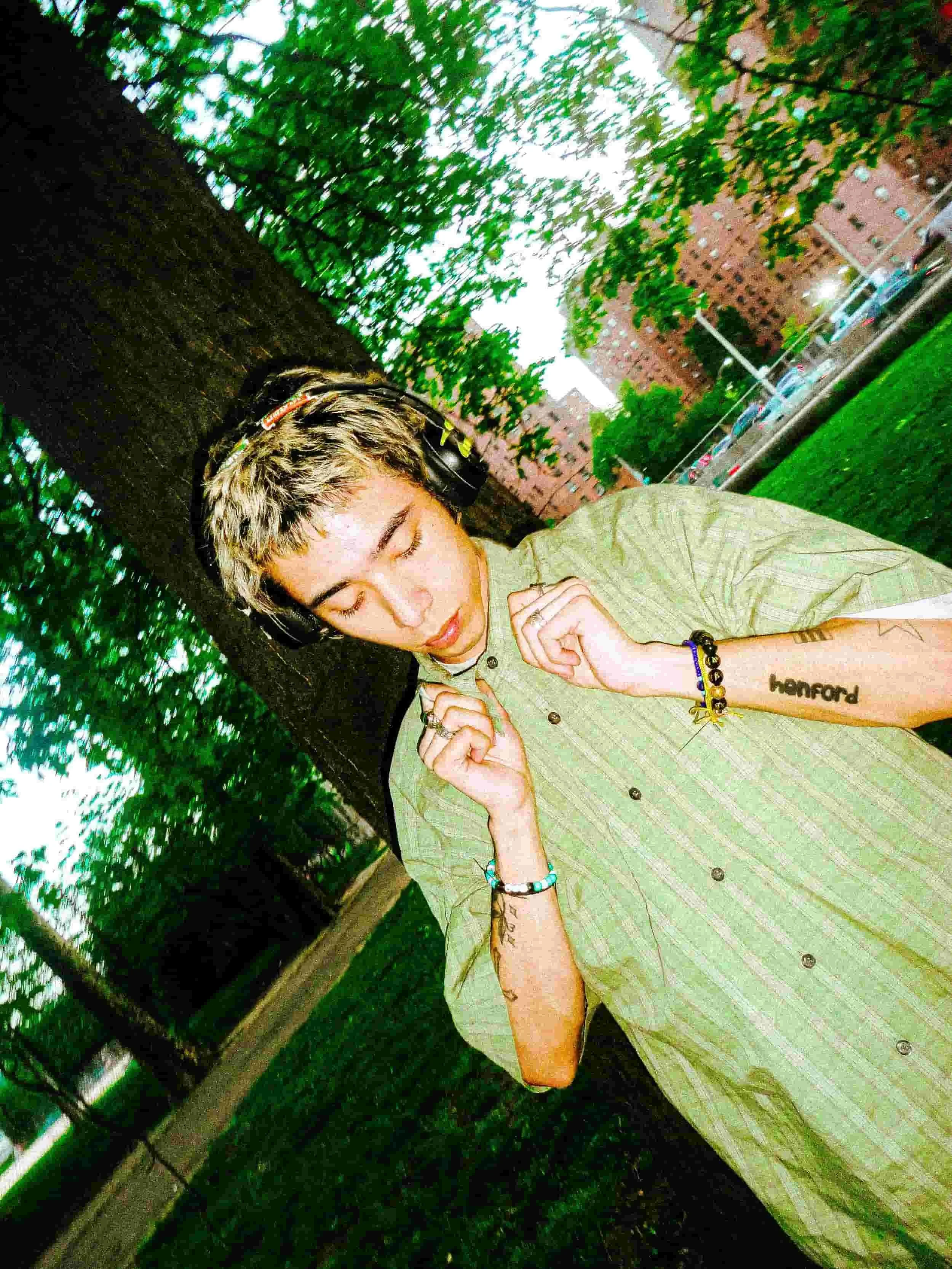 Young man with colorful hair, wearing headphones and a green striped shirt, standing outdoors near a large tree in a park with residential buildings in the background.