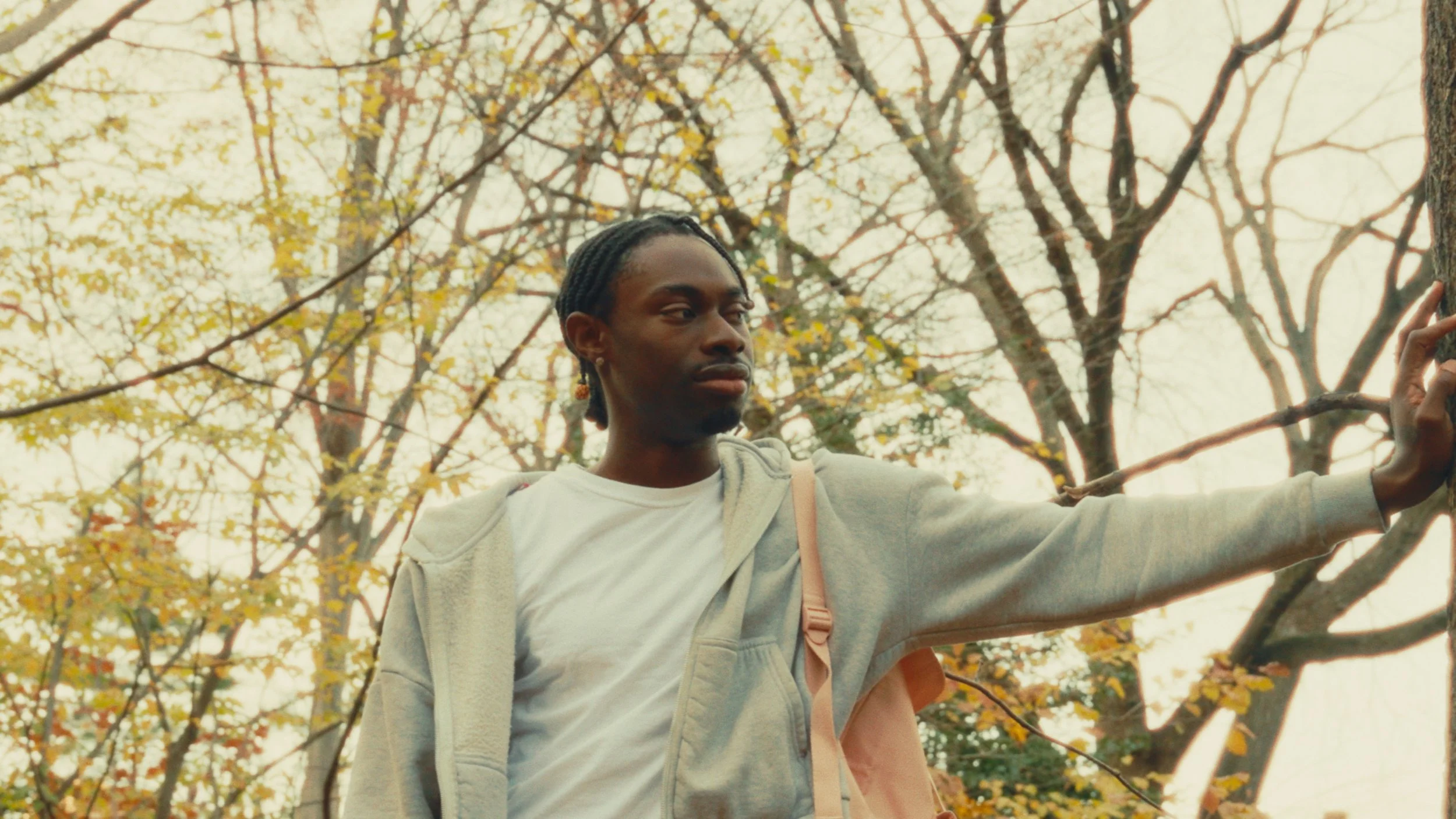 Person in white t-shirt and gray jacket standing outdoors among trees with autumn foliage, touching a tree trunk.