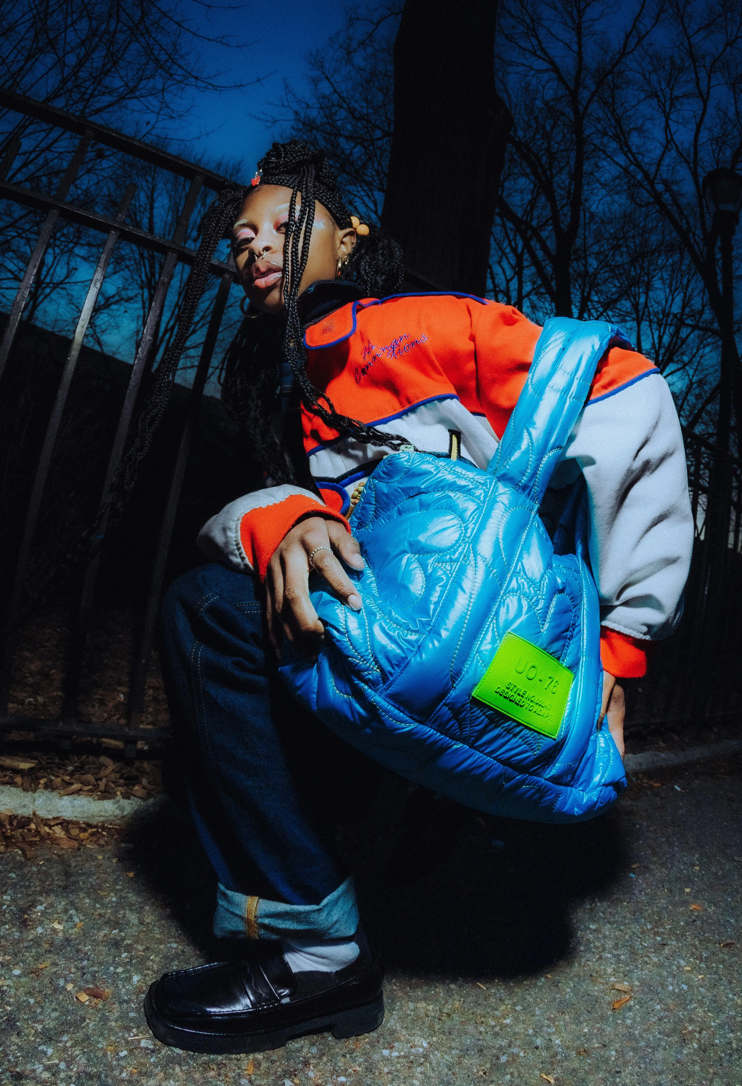 A young woman with braided hair, colorful makeup, and jewelry squats outdoors during dusk or night, holding a bright blue puffer bag, with a black metal fence and leafless trees in the background.