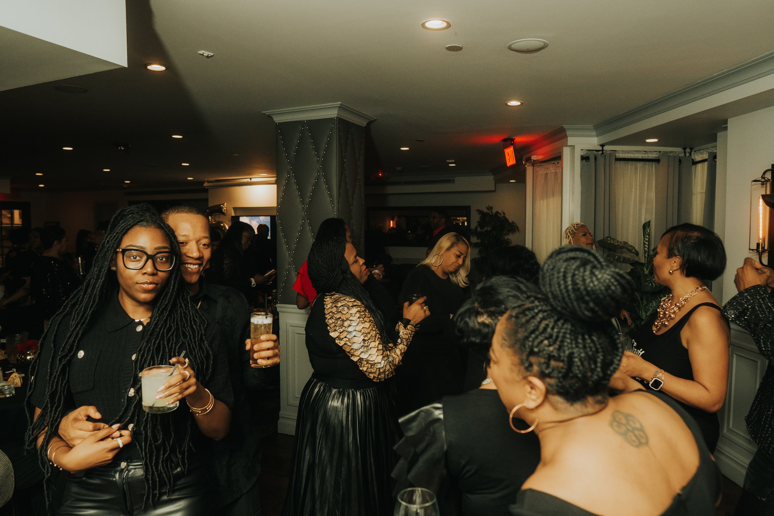 A group of people socializing at a party in a dimly lit room, with some holding drinks. The atmosphere is lively, with guests wearing fashionable attire. The setting appears to be a sophisticated indoor event space.