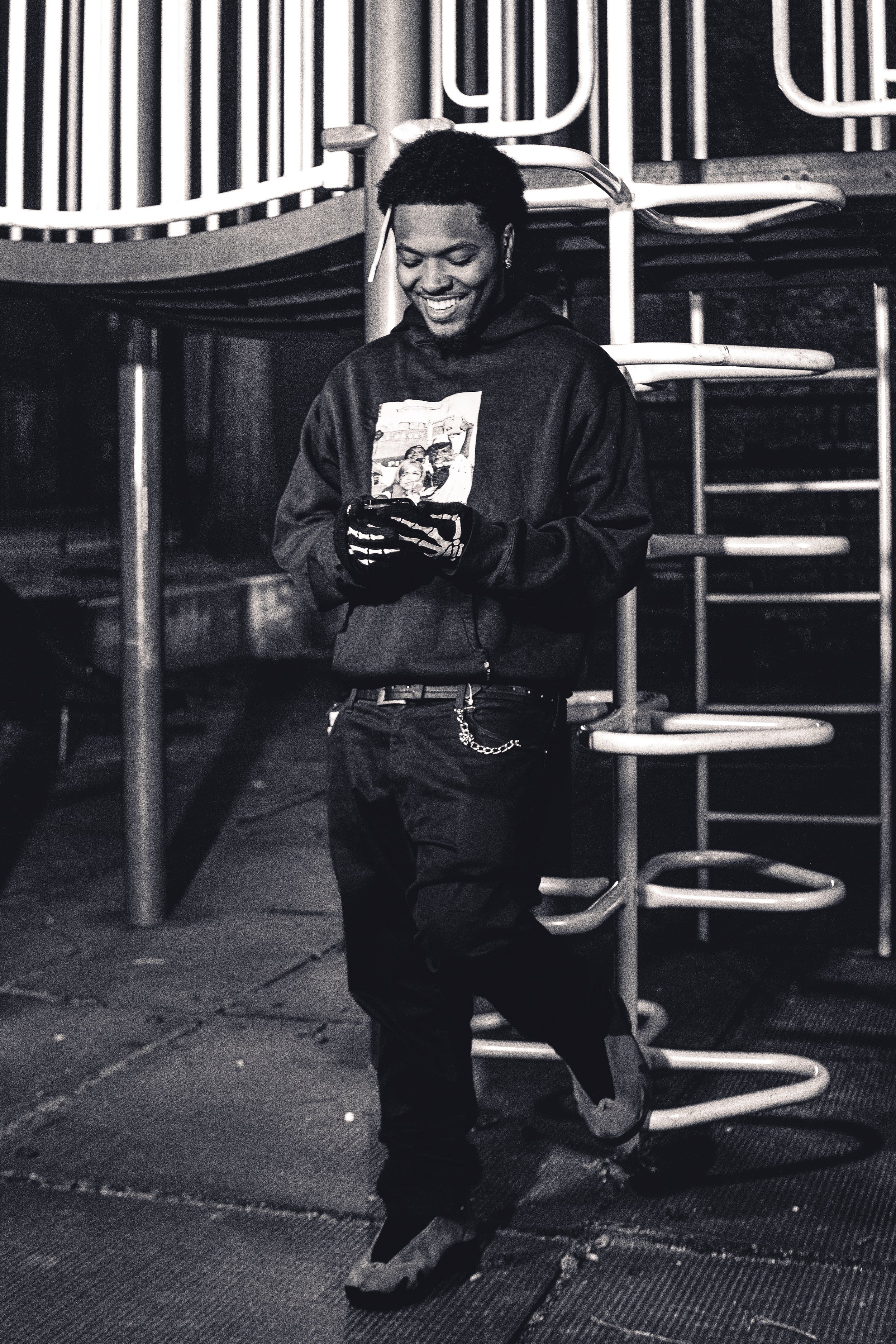 A young man smiling while looking at his phone, standing on a playground at night.