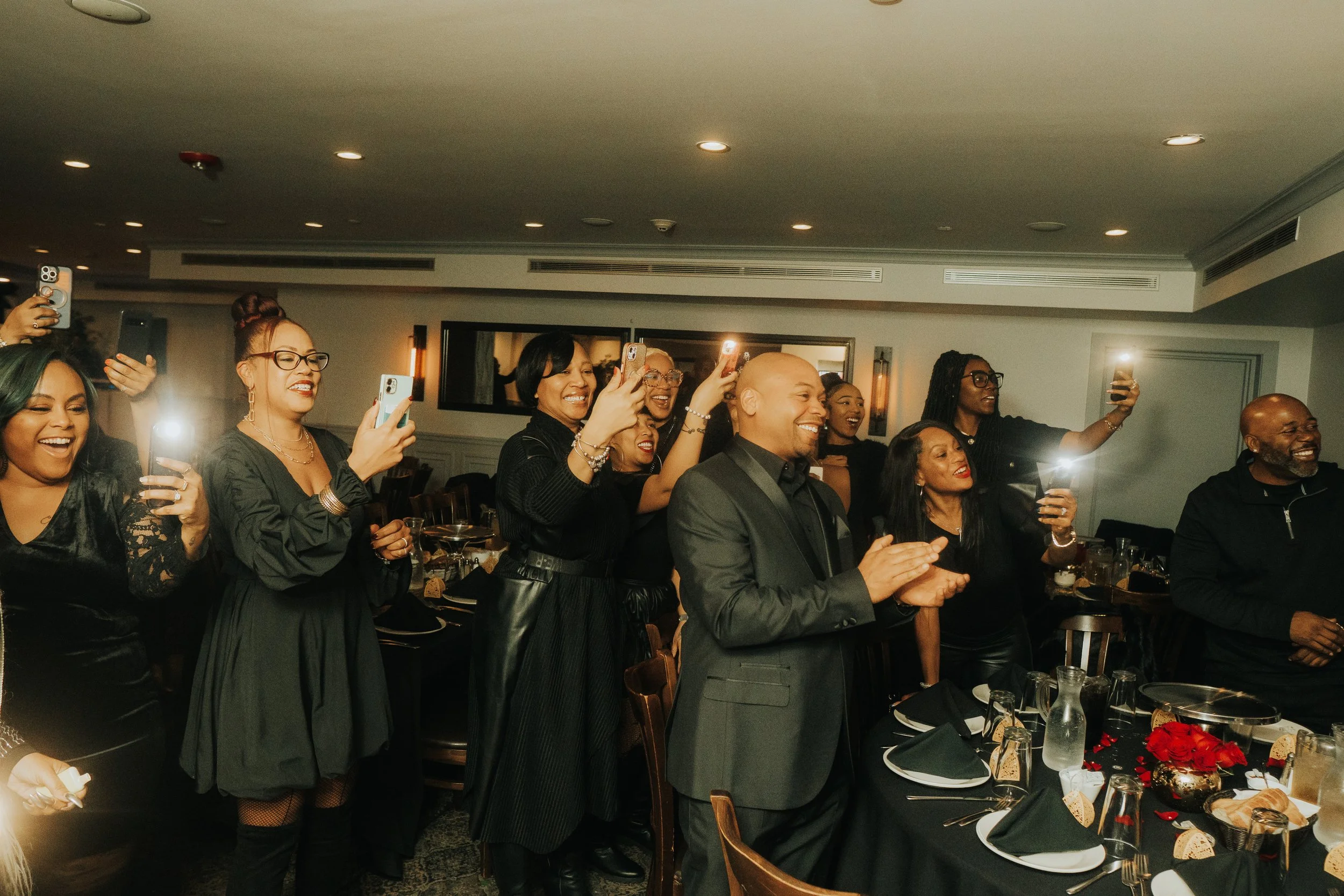 People at a celebration, smiling and taking photos with phones, wearing black outfits.