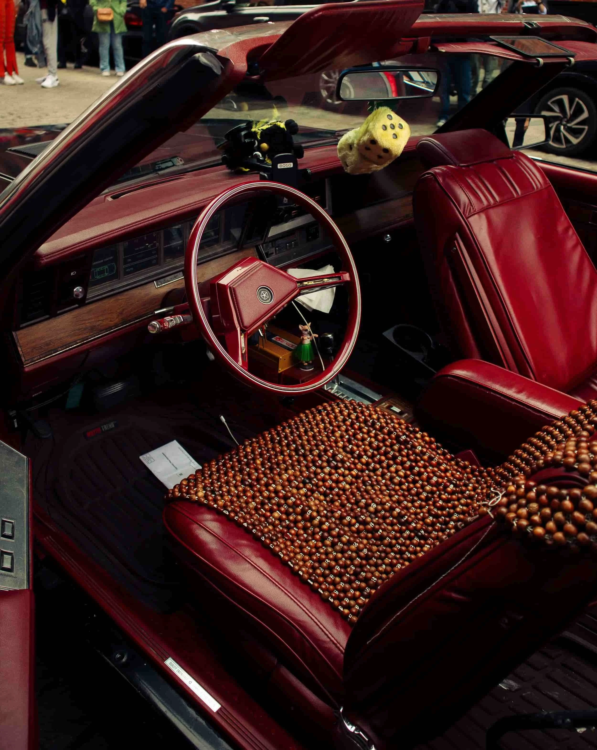 Interior of a vintage car with red leather seats, a wooden dashboard, a beaded seat cover, and fuzzy dice hanging from the rearview mirror.