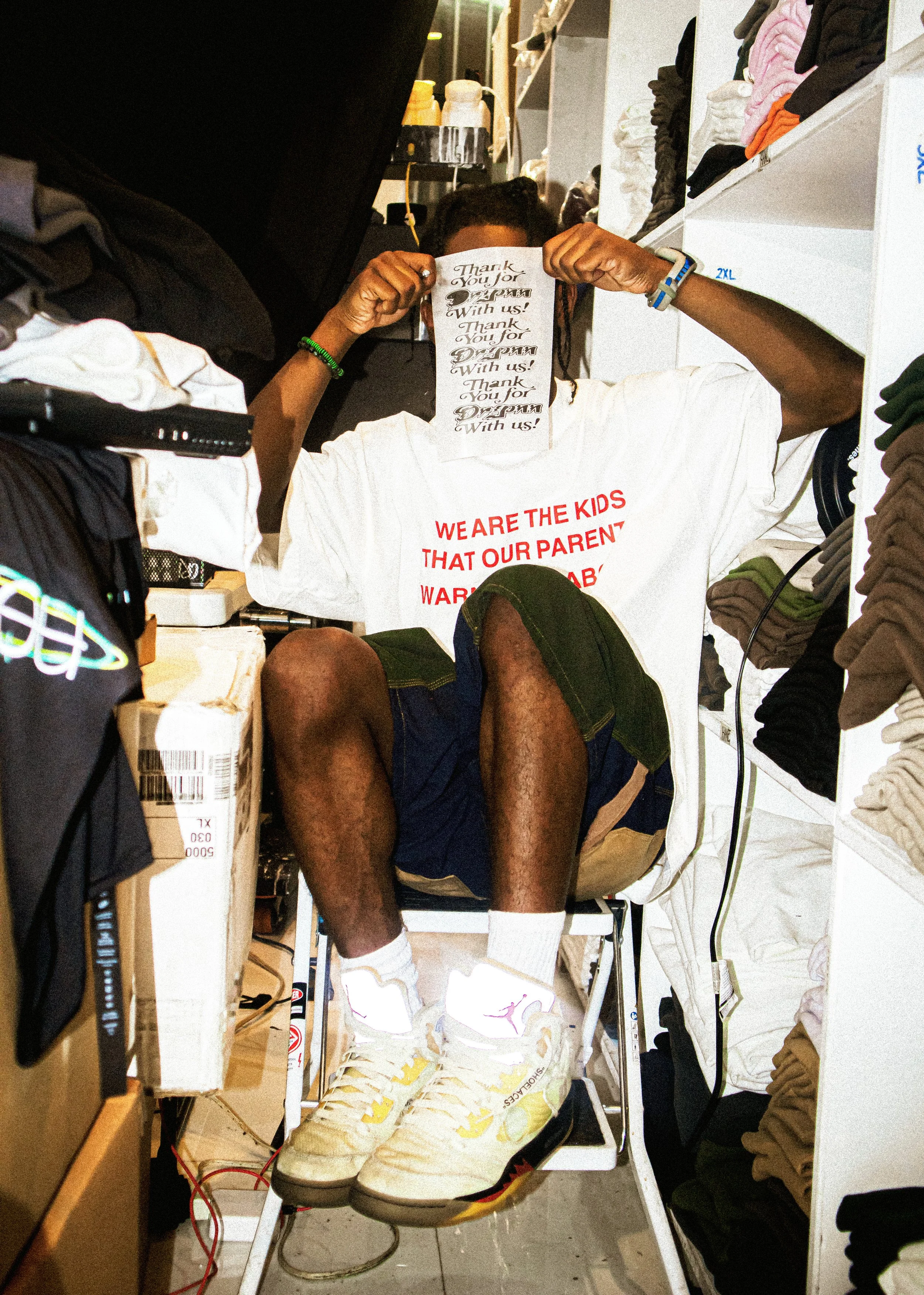 A person sitting in a small, cluttered retail or storage space, holding a paper in front of their face. They are wearing a white T-shirt with red text, dark shorts, and white sneakers with yellow accents. The space is filled with shelves of folded cl