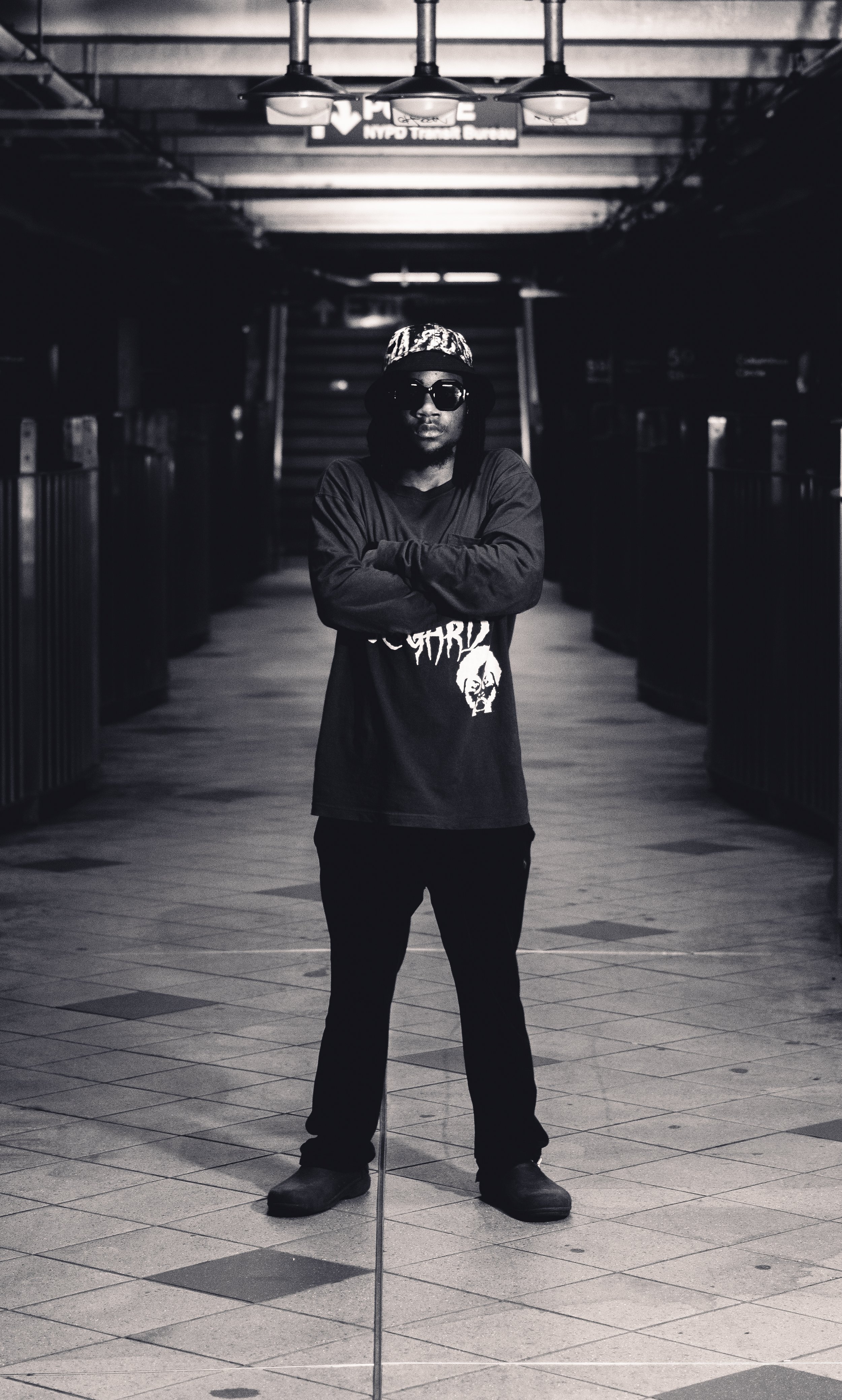 A person standing with arms crossed in an underground station or mall, wearing sunglasses, a patterned bucket hat, a black long-sleeve shirt with graphic print, black pants, and boots, in black and white photography.