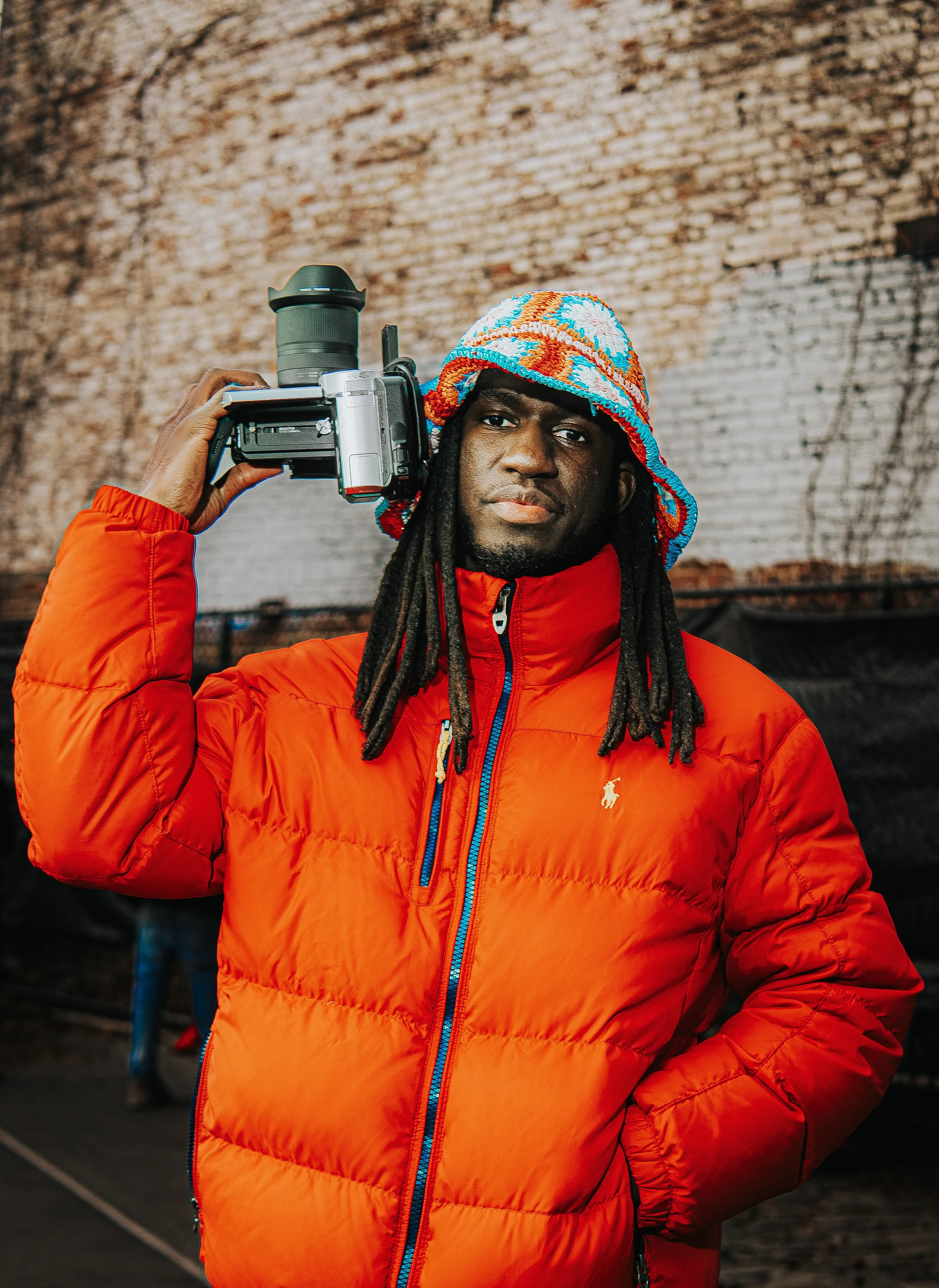 A man with dreadlocks wearing an orange puffer jacket and a colorful knit hat, holding a camera on his shoulder.