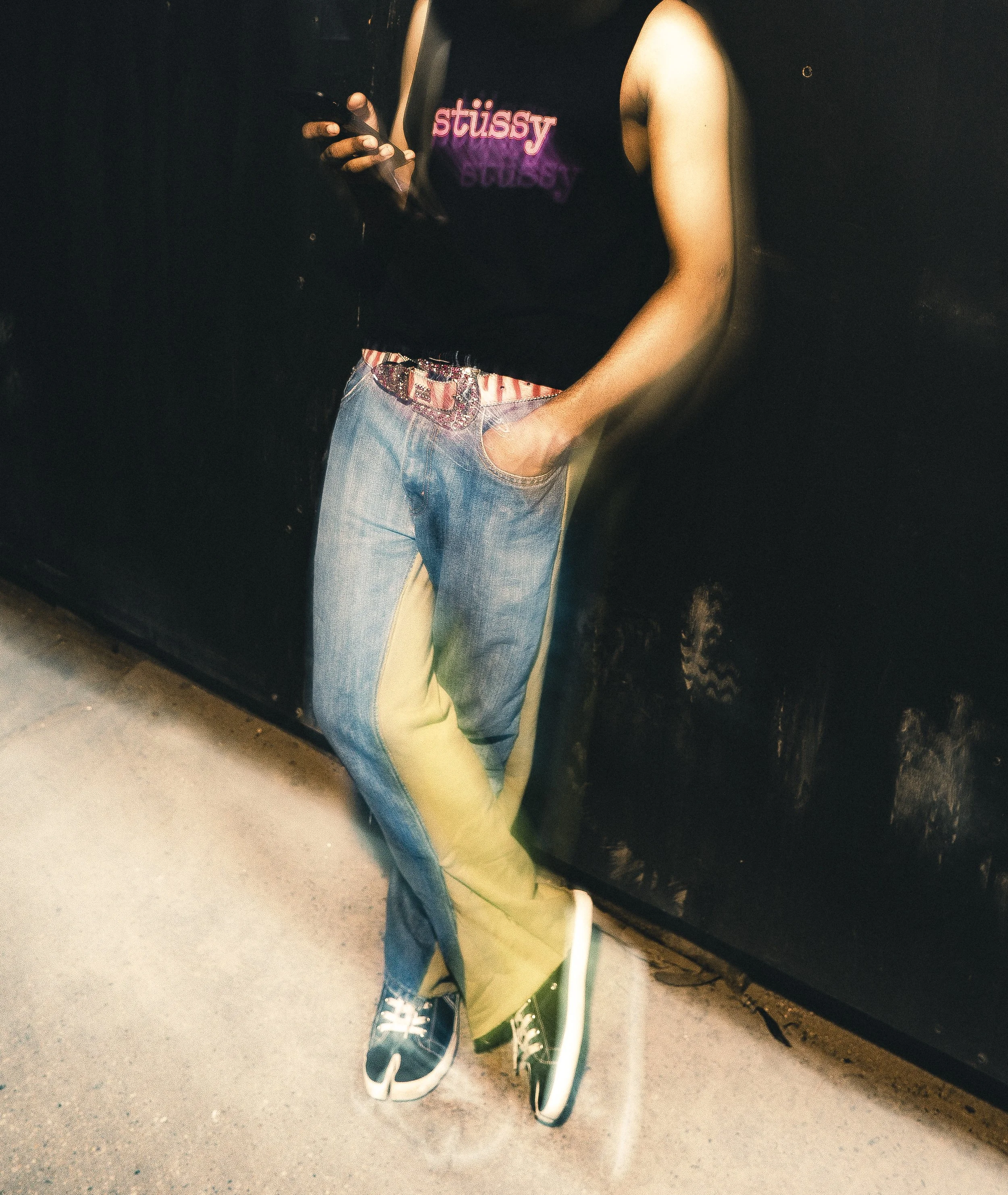 Person leaning against a black wall, wearing a black sleeveless top with pink and purple 'Stussy' logo, colorful jeans with yellow and blue sections, and dark sneakers with white soles.