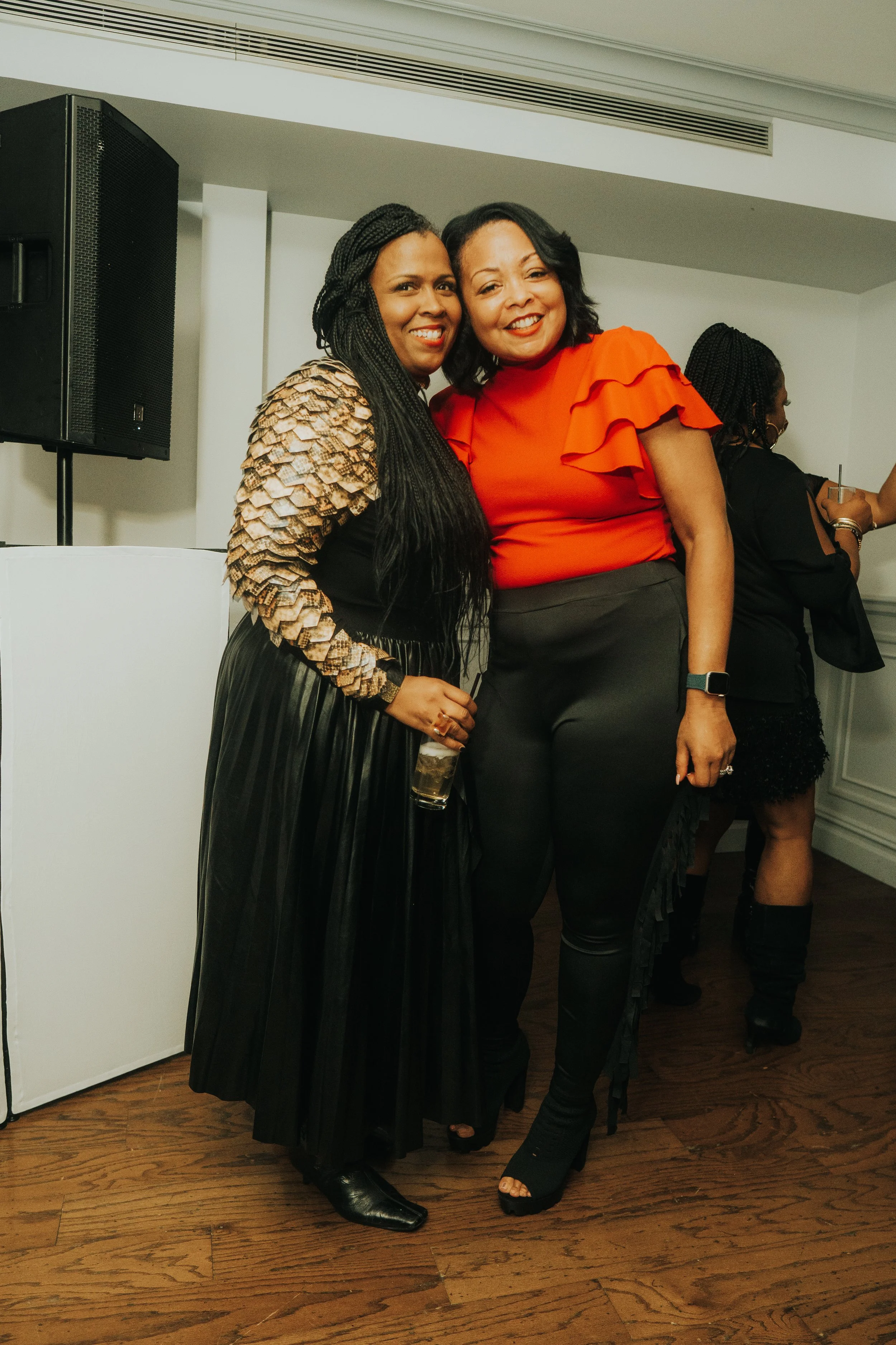 Two women standing together, one wearing a black and gold outfit, the other in a red top, in a room with wood flooring.