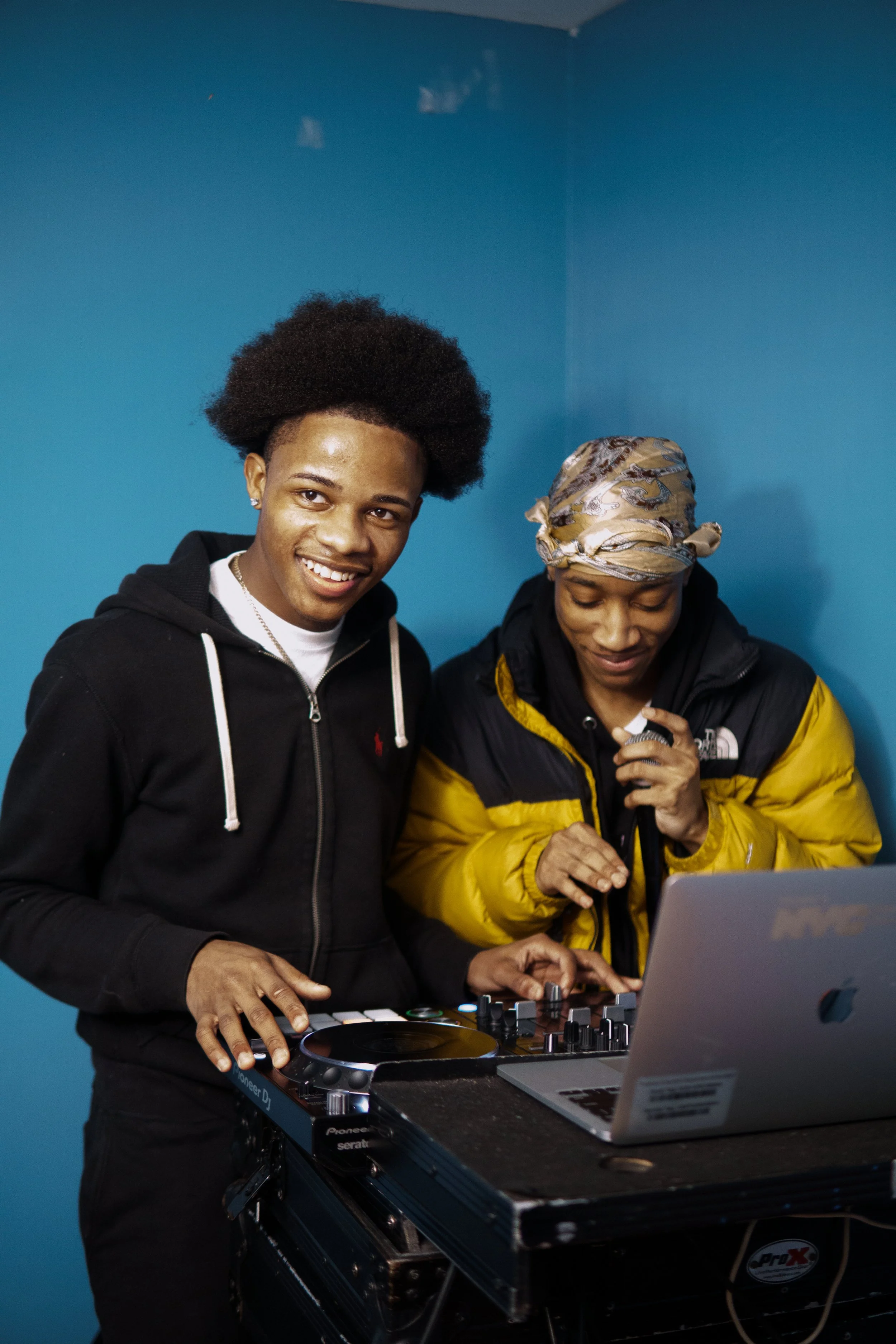 Two young men are using DJ equipment and a laptop against a blue wall. One is smiling at the camera while the other is focused on the equipment.
