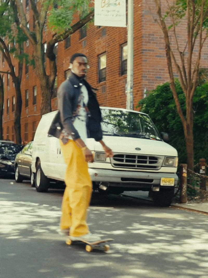 A man skateboarding on a city street with brick apartment buildings and parked cars, including a white van, in the background.