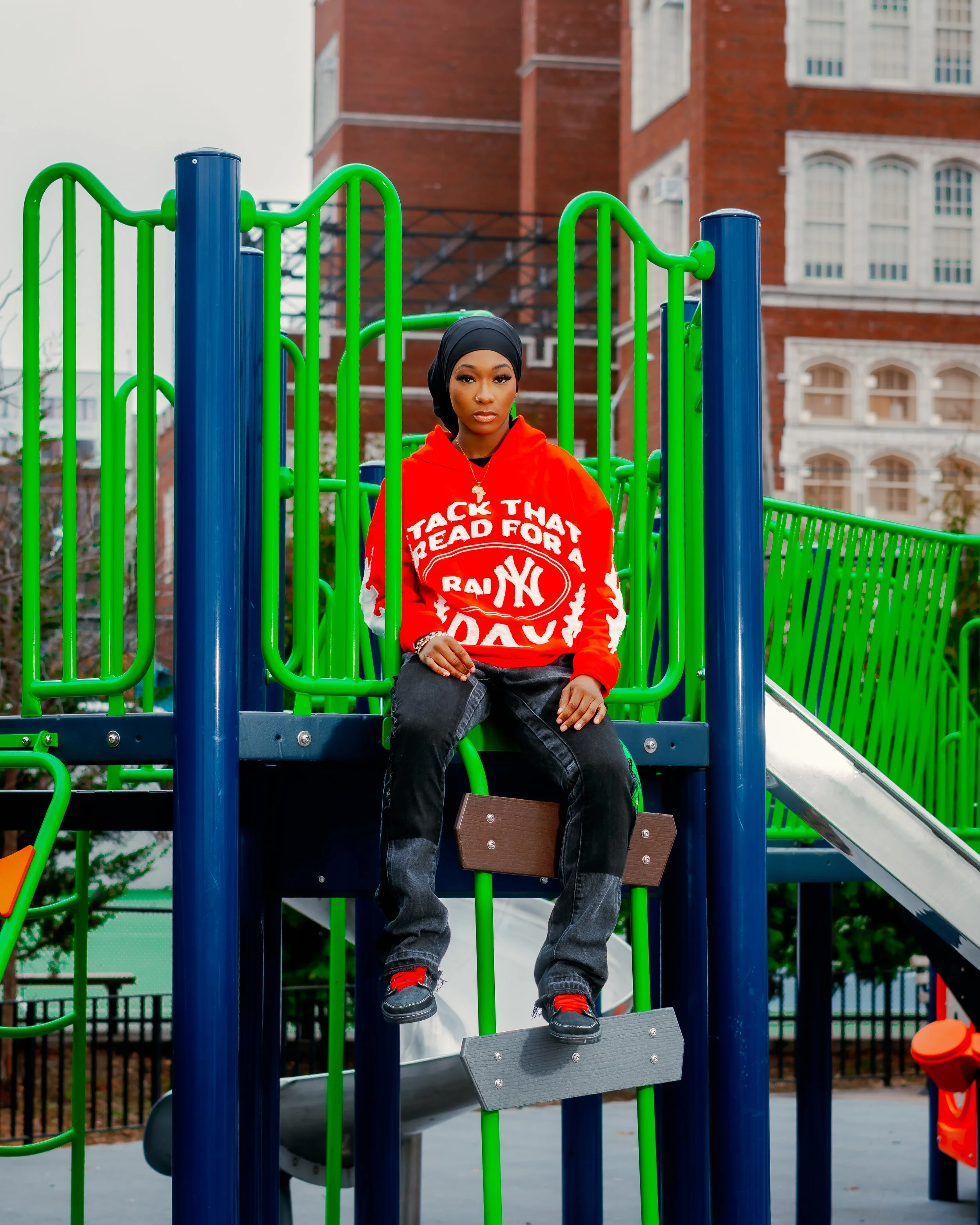 A woman in a black headscarf and red hoodie sitting on a playground structure with green railings and a slide in an urban park setting, with brick buildings in the background.