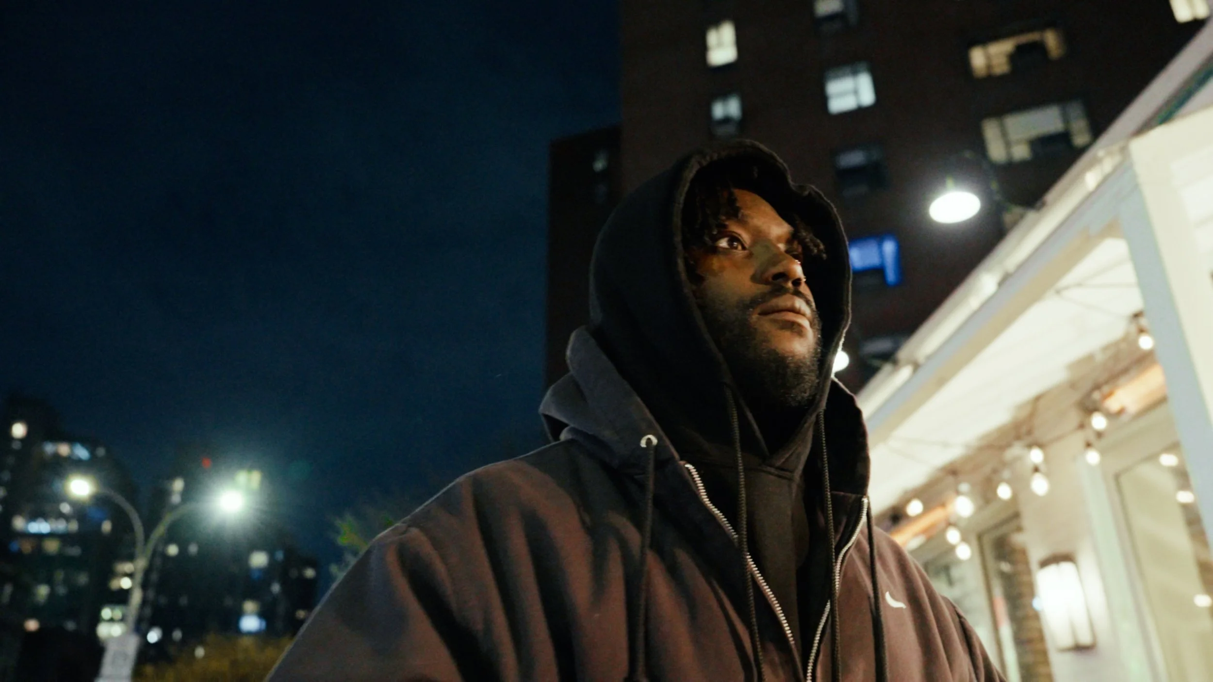 Person wearing a hoodie at night with urban buildings in the background.