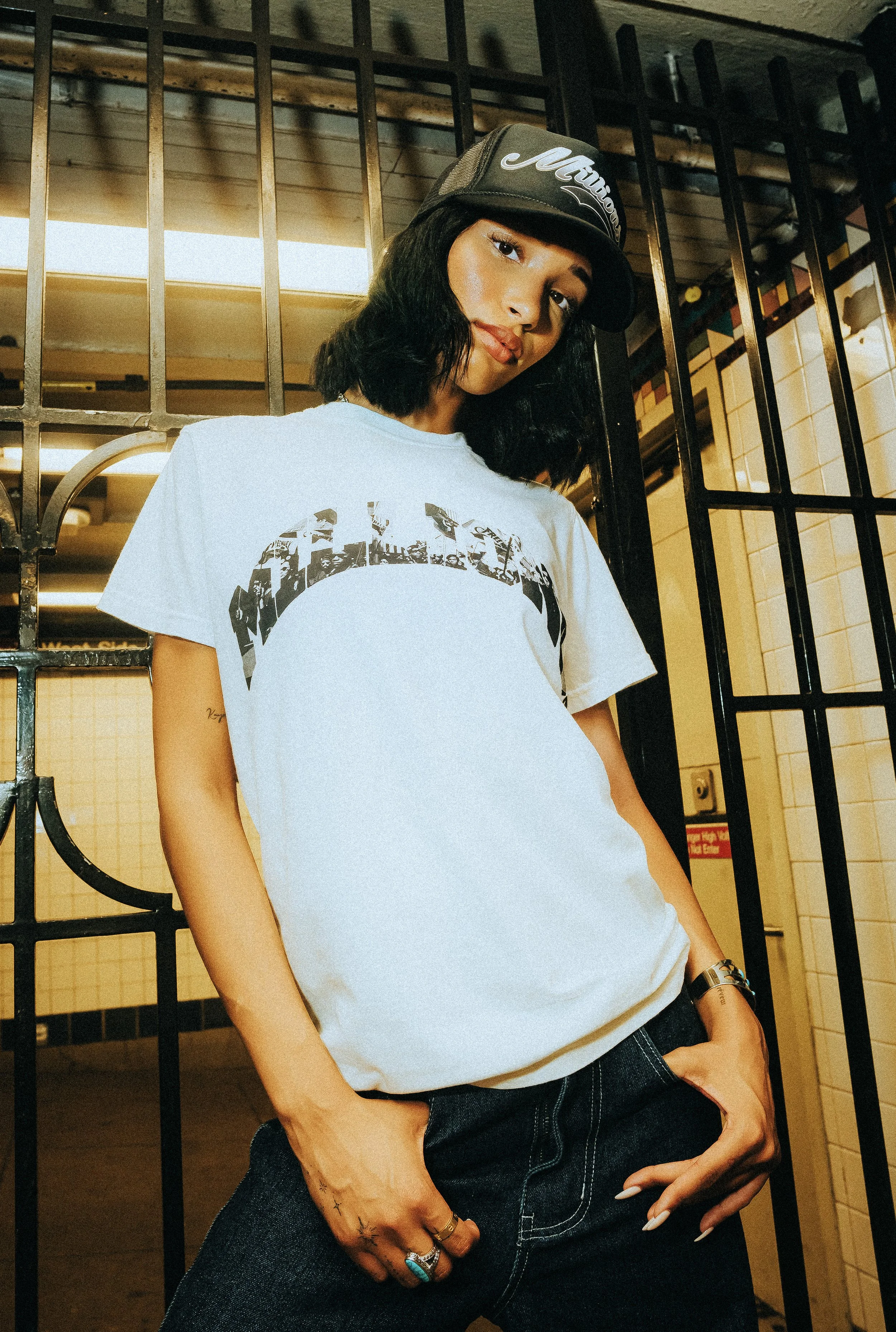 A woman with black, shoulder-length hair, wearing a black baseball cap, a white graphic T-shirt, dark jeans, and jewelry, posing in front of a metal gate and tiled wall.