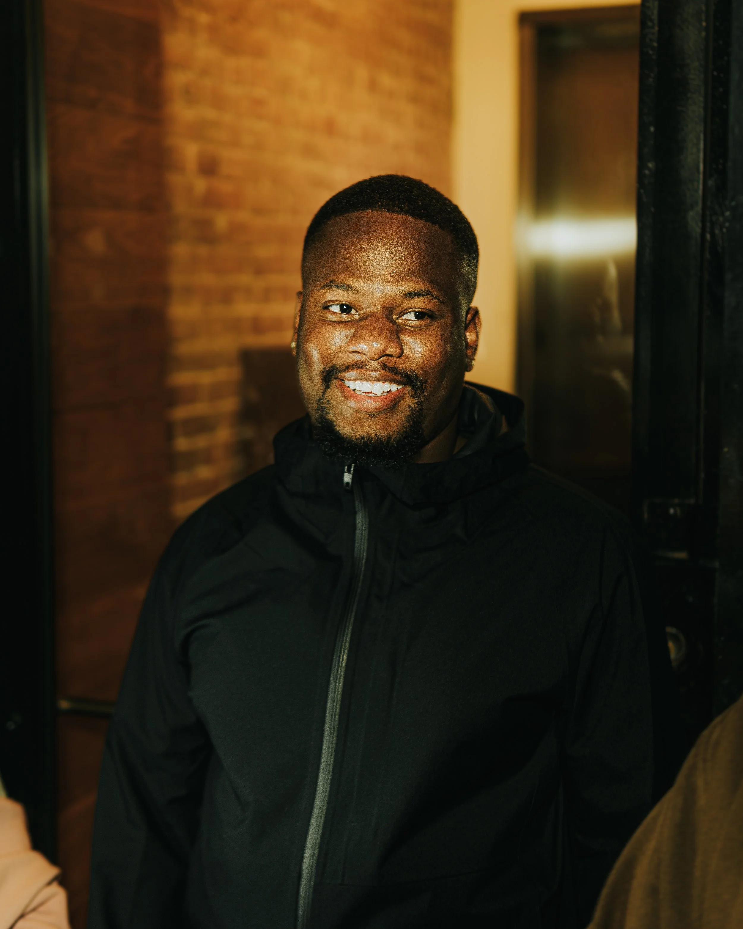 A smiling man with a short beard and black jacket standing in front of a brick wall and window at night.